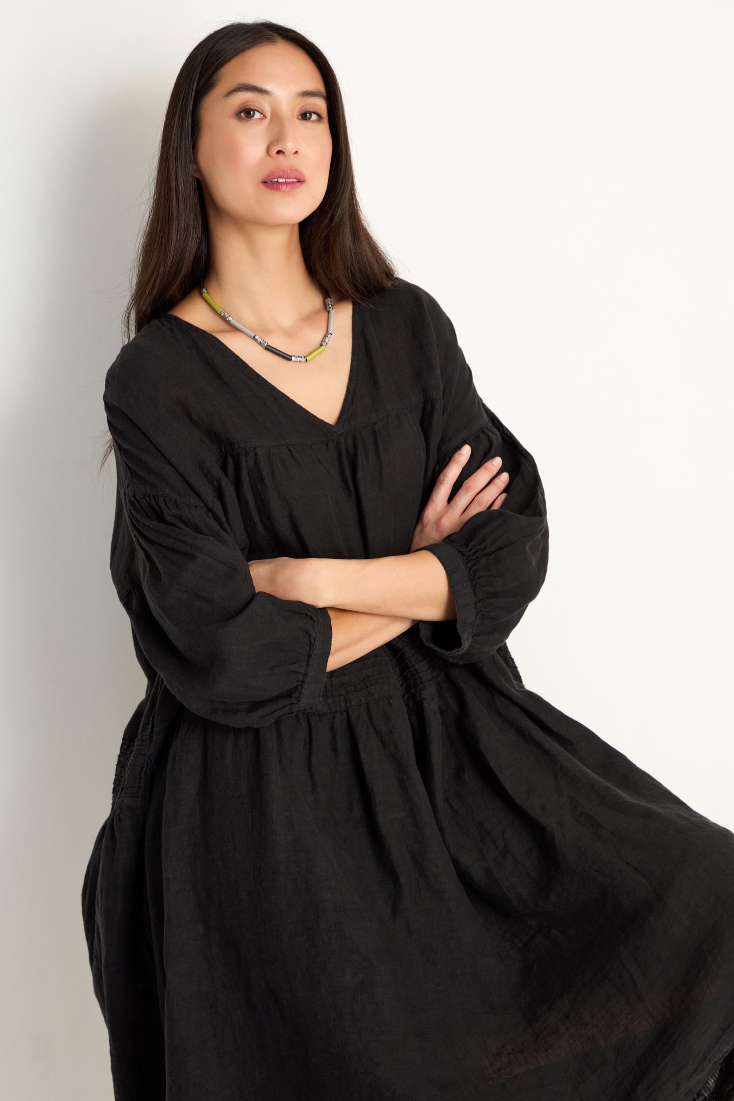 A woman with long dark hair and a necklace stands against a plain light background, arms crossed, wearing the Easy Fit V-Neck Black Linen Dress.