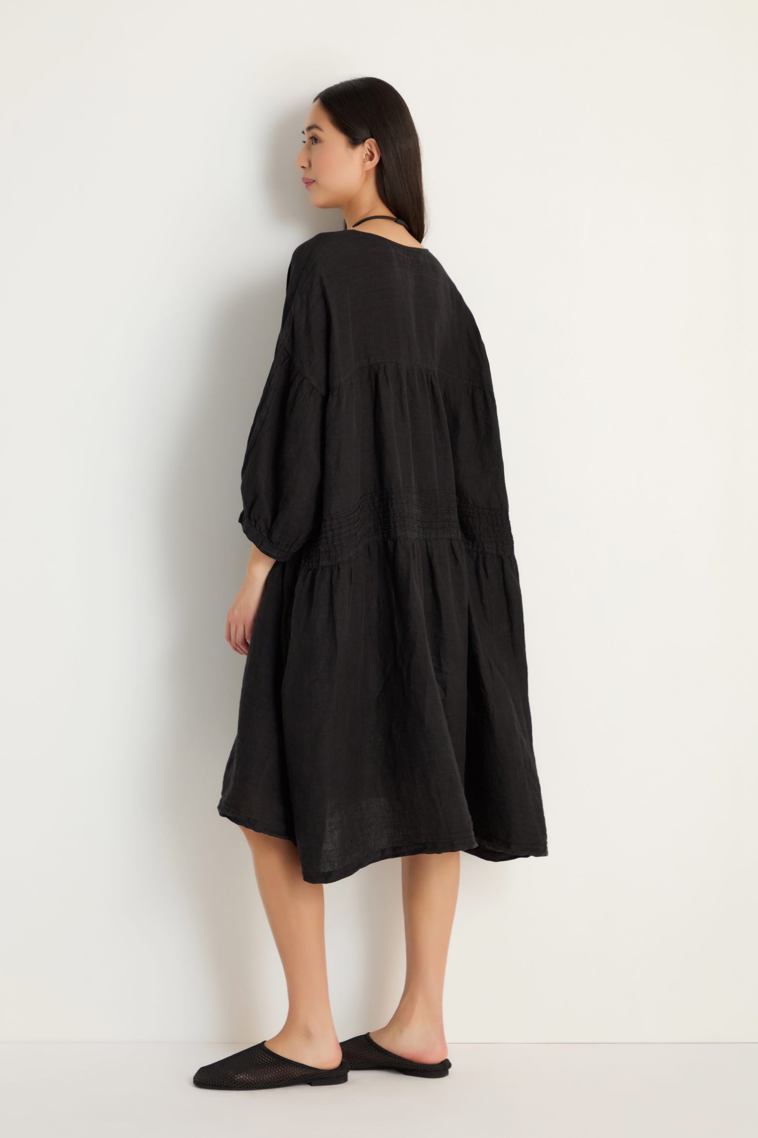 A woman stands slightly turned from the camera, wearing the Easy Fit V-Neck Black Linen Dress with black slip-on shoes, set against a plain white background.