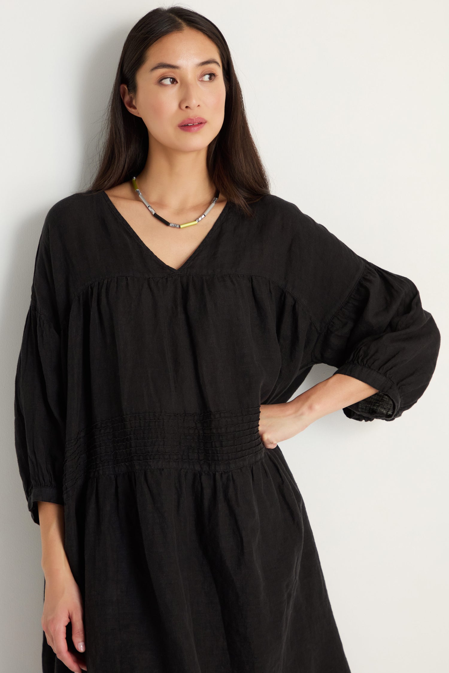 A woman with long dark hair wears the Easy Fit V-Neck Black Linen Dress and a beaded necklace as she stands against a plain light-colored wall.