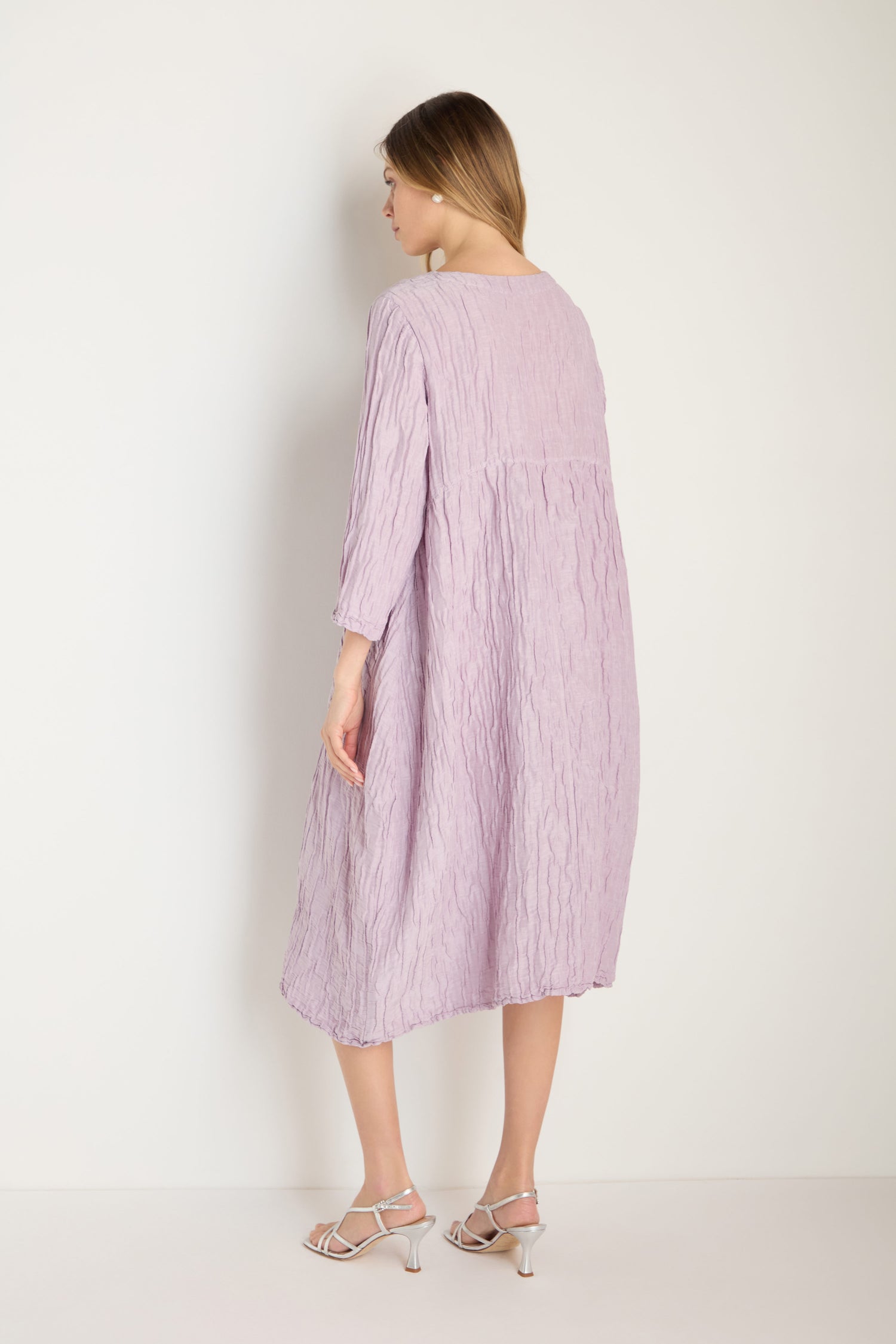 A woman faces away, wearing a loose Silk Linen Lilac Patch Pocket Dress with three-quarter sleeves and white heeled sandals, set against a plain white background.
