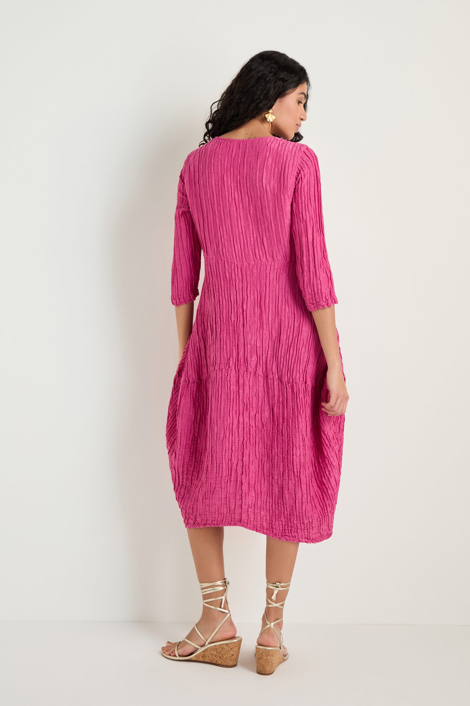 A woman stands facing away, wearing the Silk Linen Bubble Dress in fuchsia with three-quarter sleeves and knee-length textured weave, paired with beige wedge sandals.