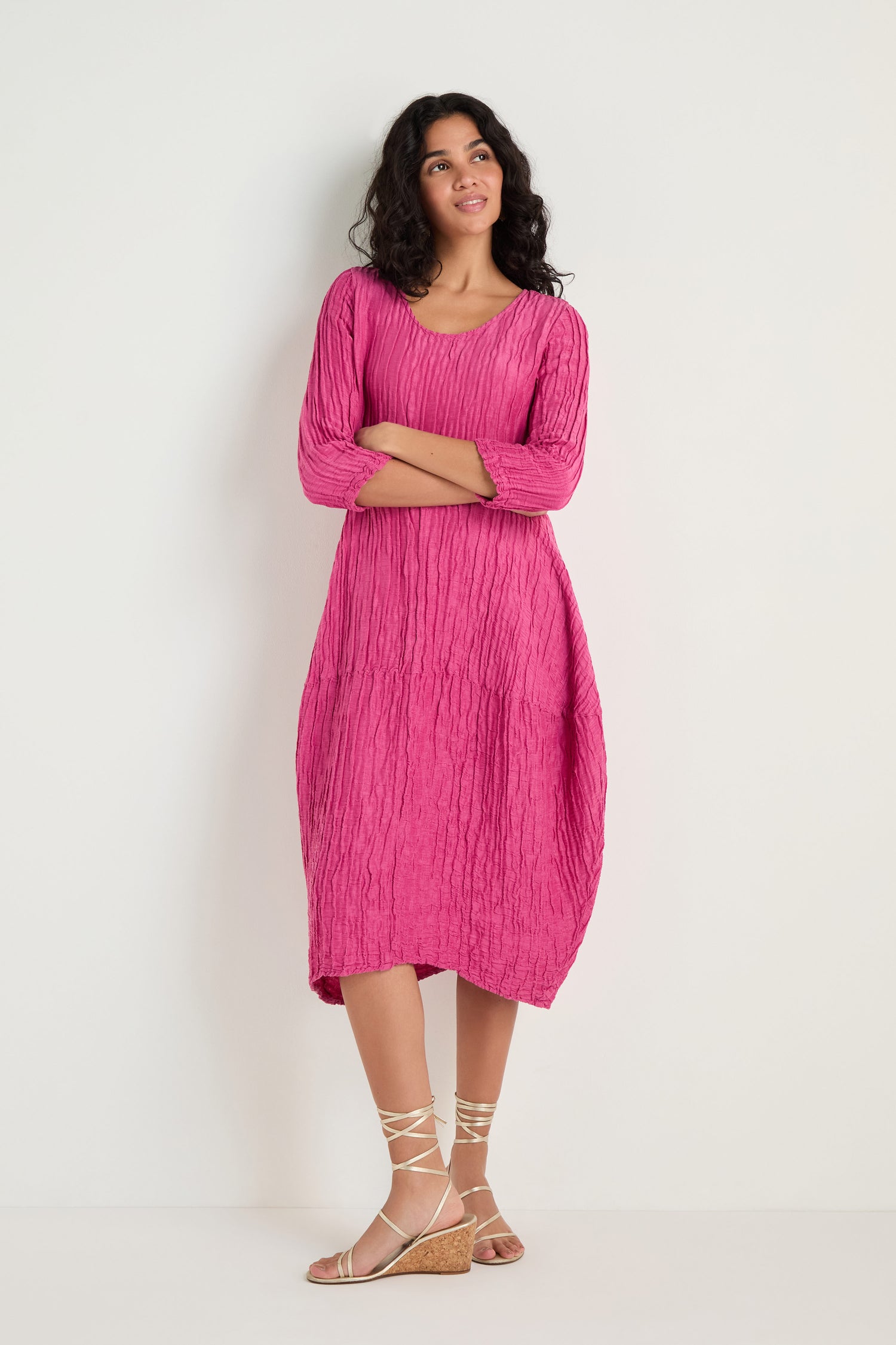 A woman stands against a white background wearing the Silk Linen Bubble Dress in fuchsia, featuring a textured weave and three-quarter sleeves, paired with beige lace-up wedge sandals.
