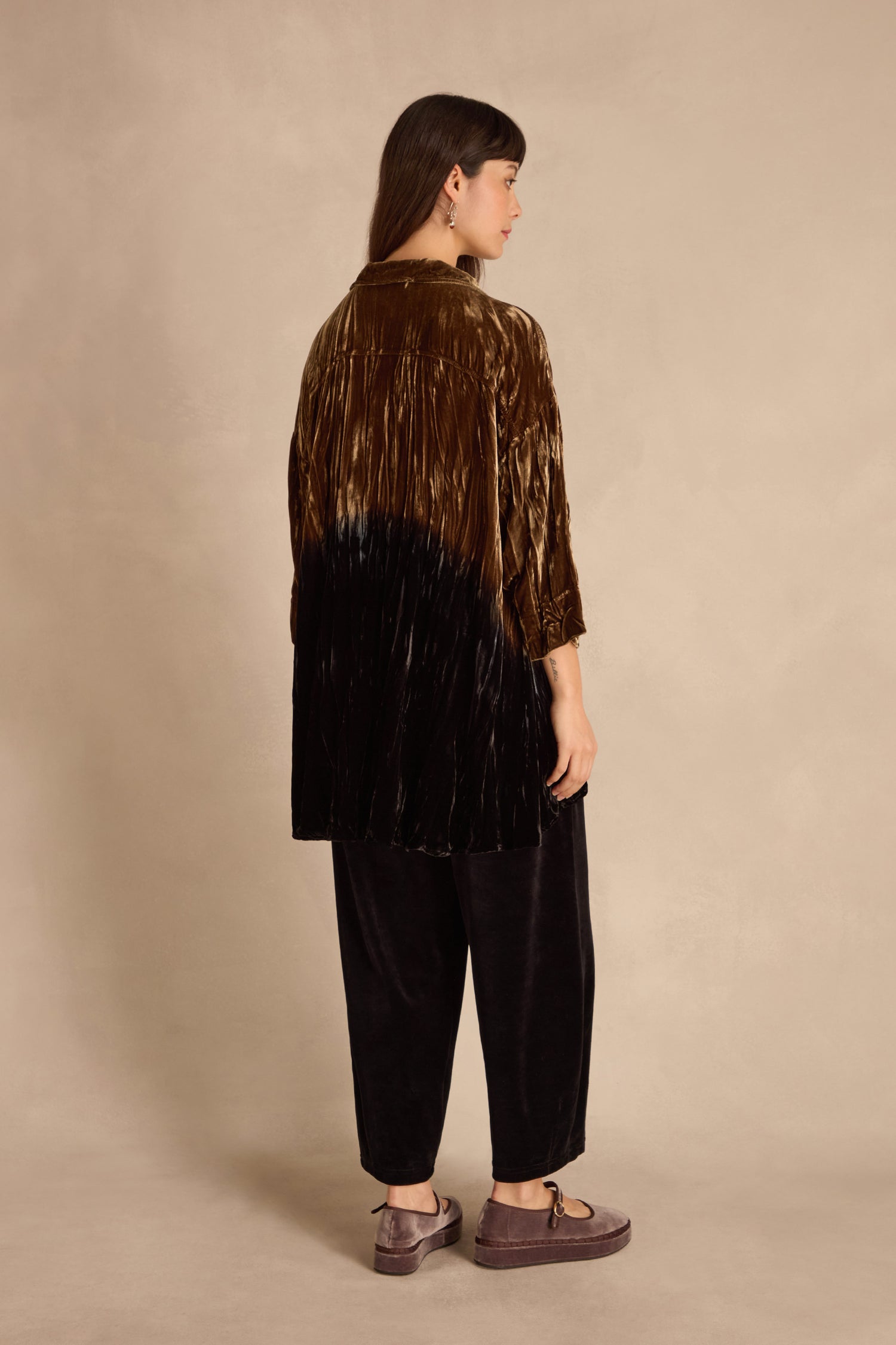 Woman stands sideways against a beige background in an Olive Silk Velvet Ombre Shirt, black velvet pants, and brown platform shoes. Her straight hair falls past her shoulders.