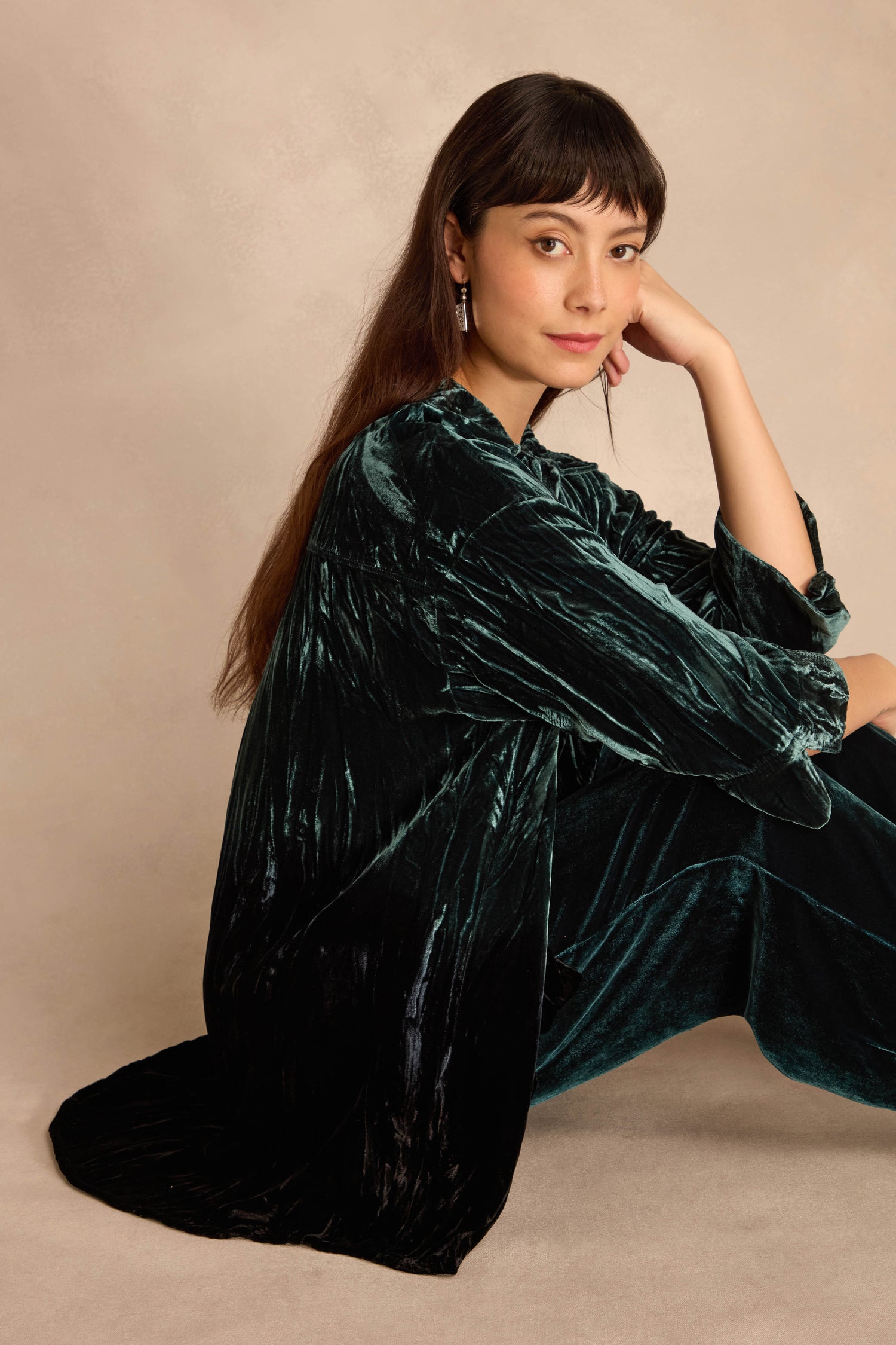 Woman with long dark hair sits on the floor, wearing the Turquoise Silk Velvet Ombre Shirt, resting her head on her hand and looking at the camera against a neutral background.