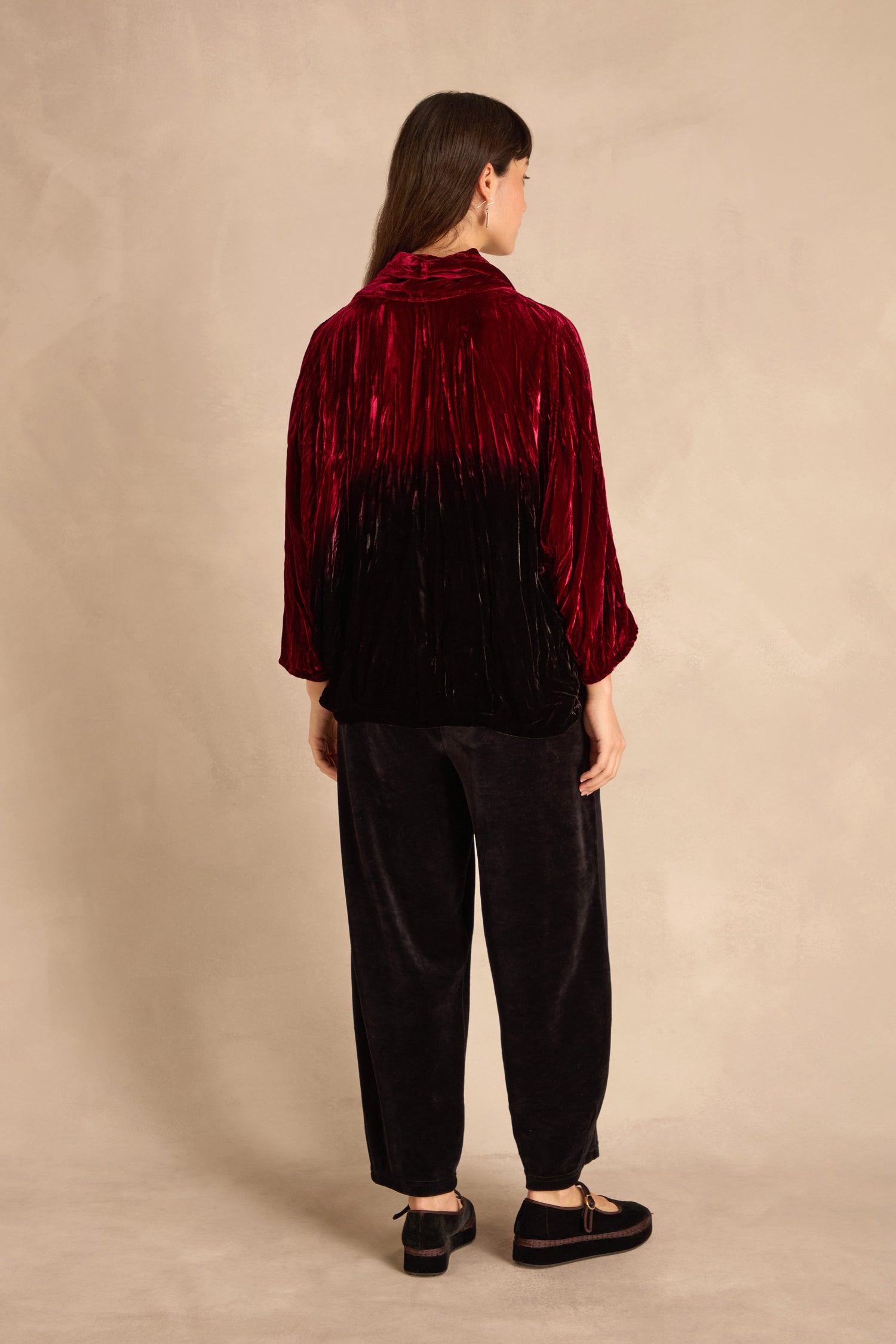 A person stands facing away, wearing the Ruby Silk Velvet Ombre Cowl Neck Top with black velvet pants and shoes, against a neutral beige background.