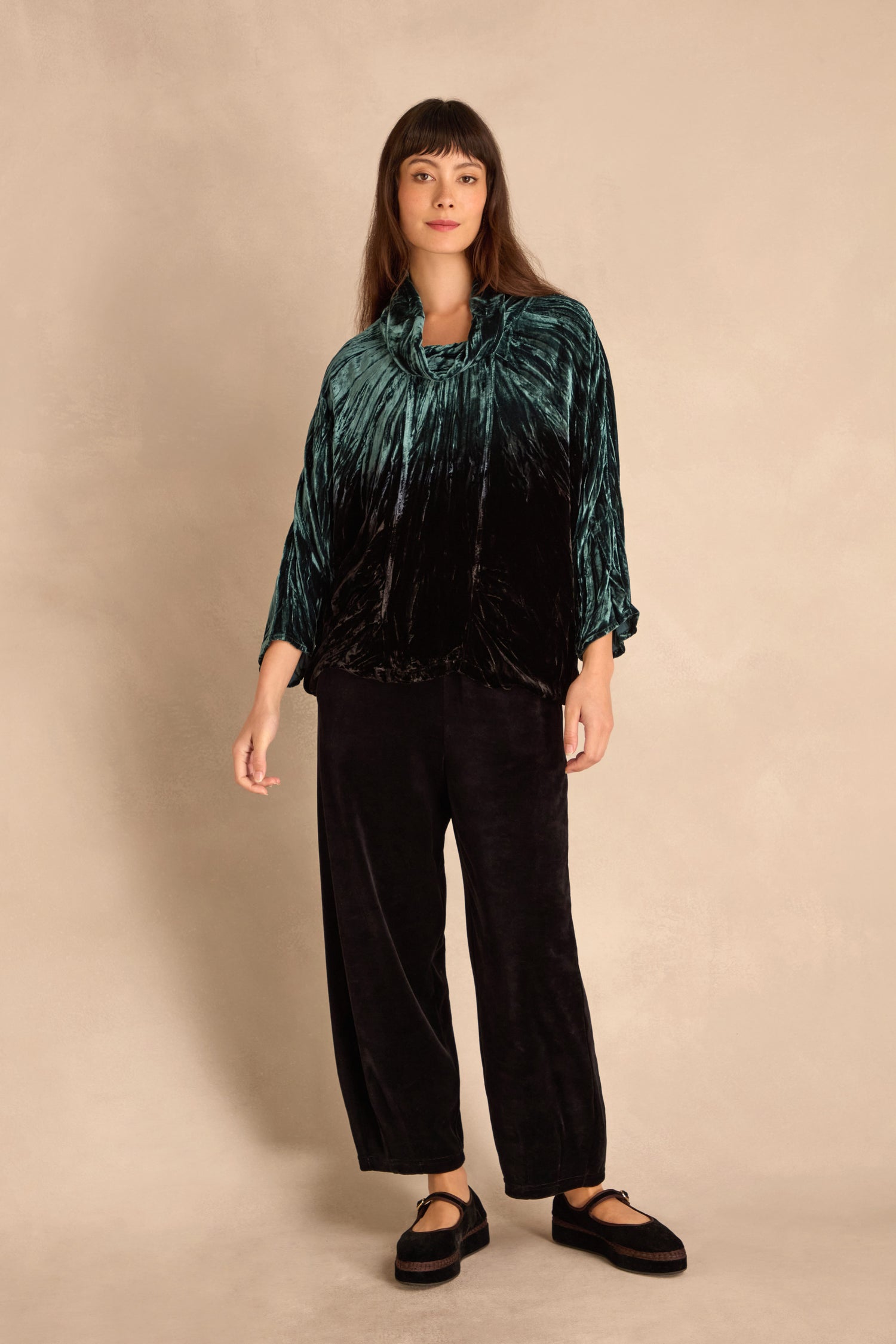 Woman standing against a beige background, wearing the Turquoise Silk Velvet Ombre Cowl Neck Top with black wide-legged pants and black platform shoes.