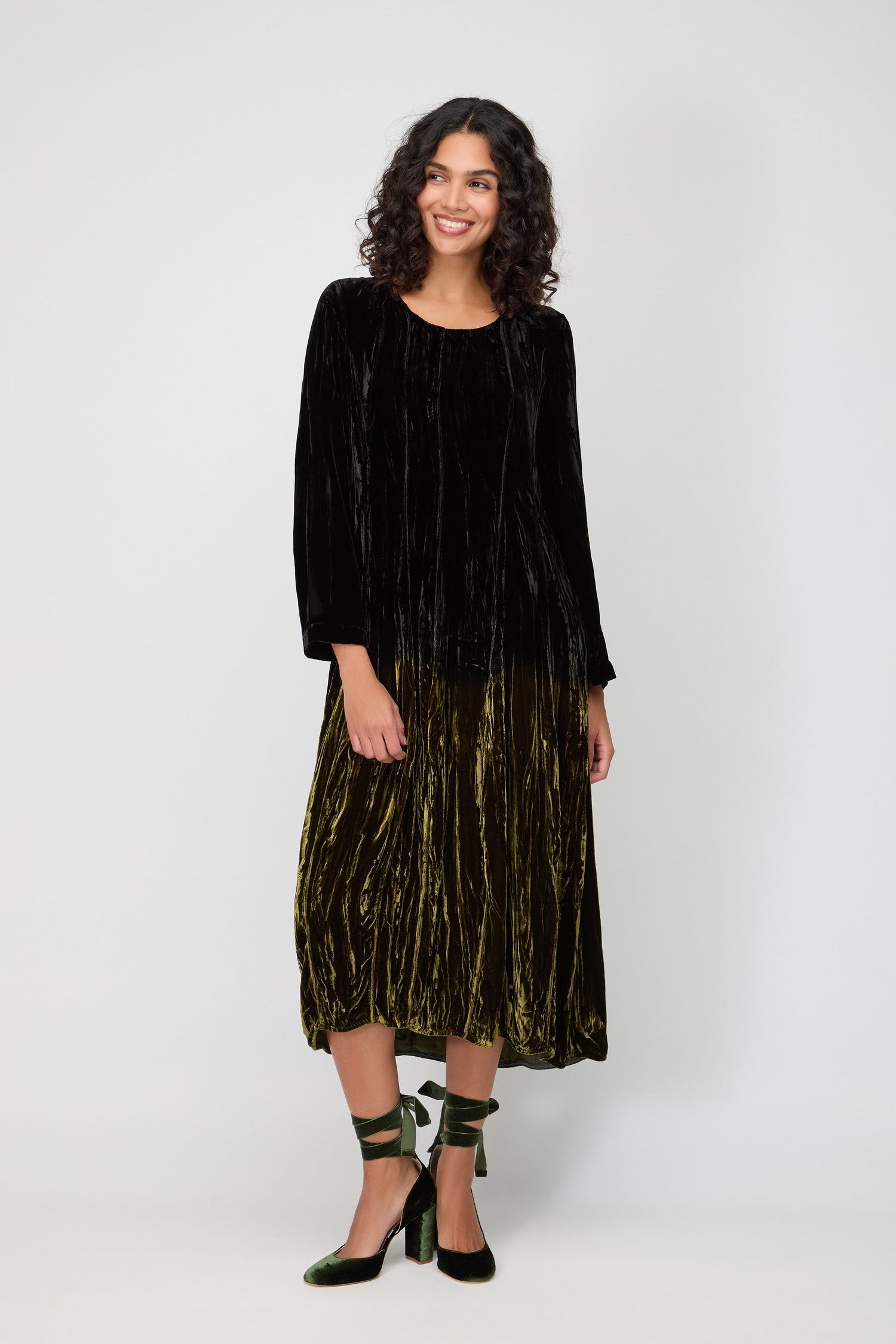 A woman stands against a plain background, wearing the Silk Velvet Ombre Dress—a long-sleeve black velvet top flowing into an olive-green skirt—paired with green ankle-tie heels.
