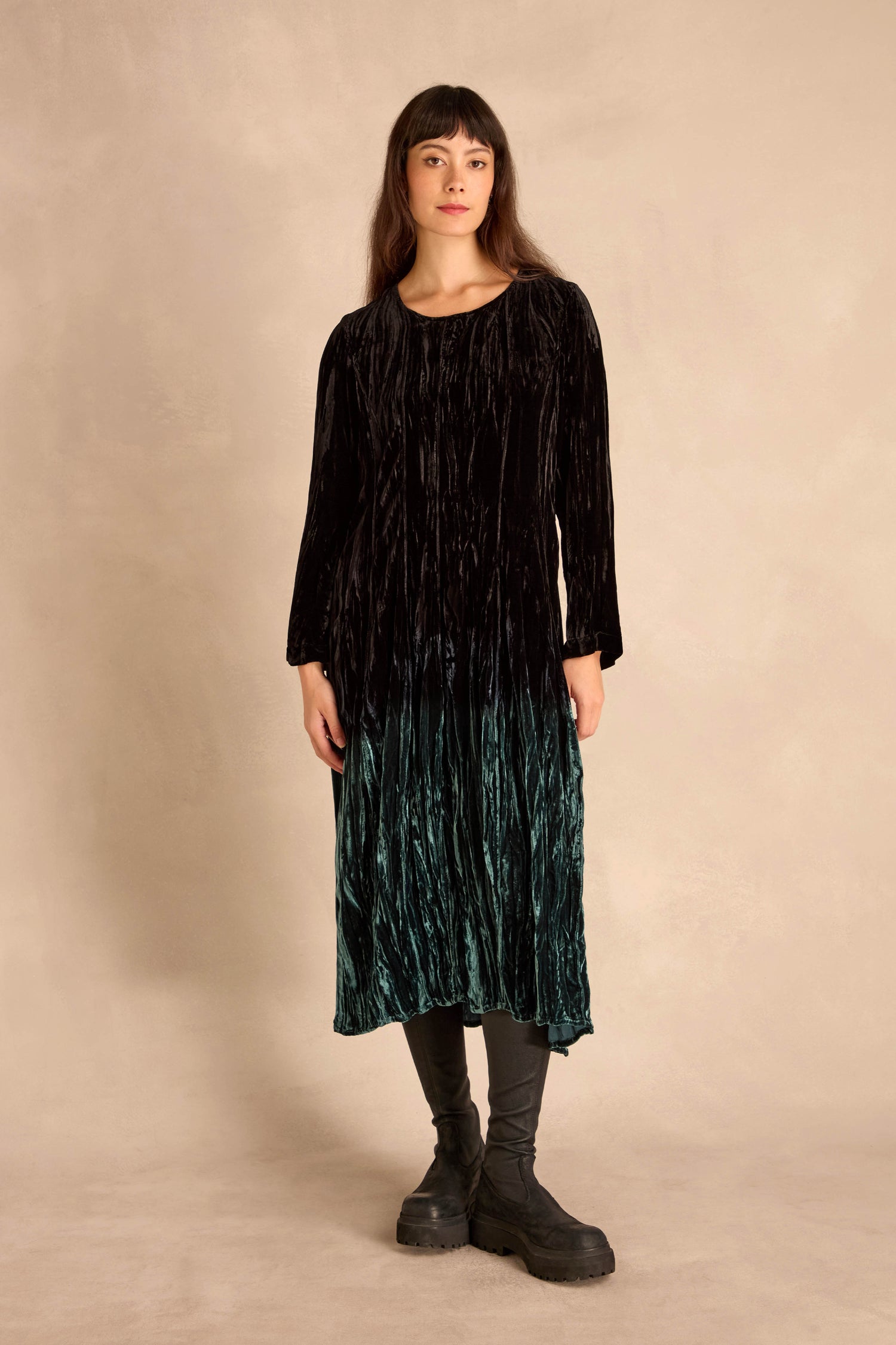 A woman stands against a beige background wearing the Silk Velvet Ombre Bubble Dress in black and green with black leggings and chunky black shoes.