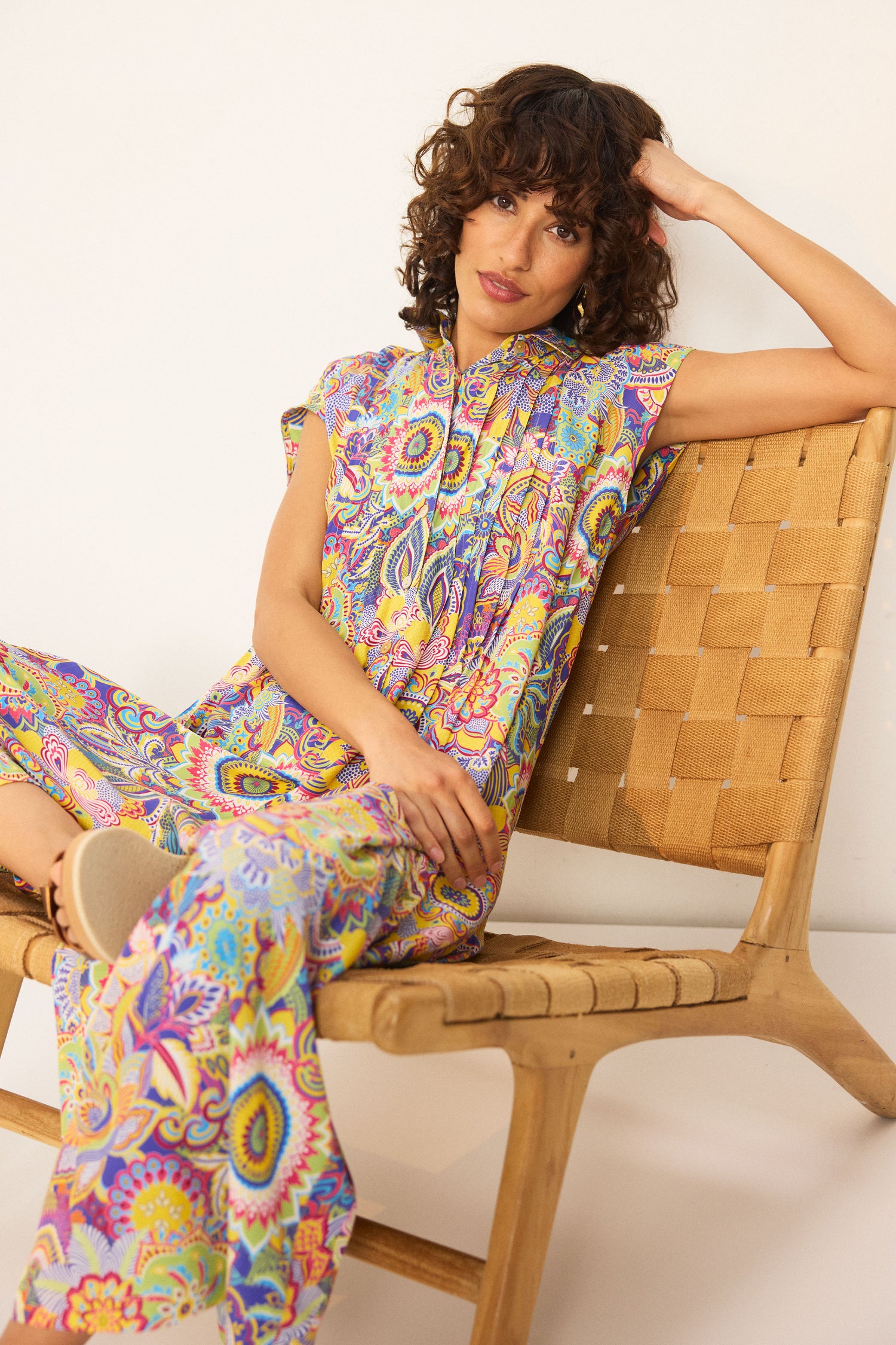 A woman with curly hair sits on a woven chair, wearing the Summer Sarasa Jumpsuit—short-sleeved, wide-leg pants in colorful patterned silk-like viscose—against a plain white background.