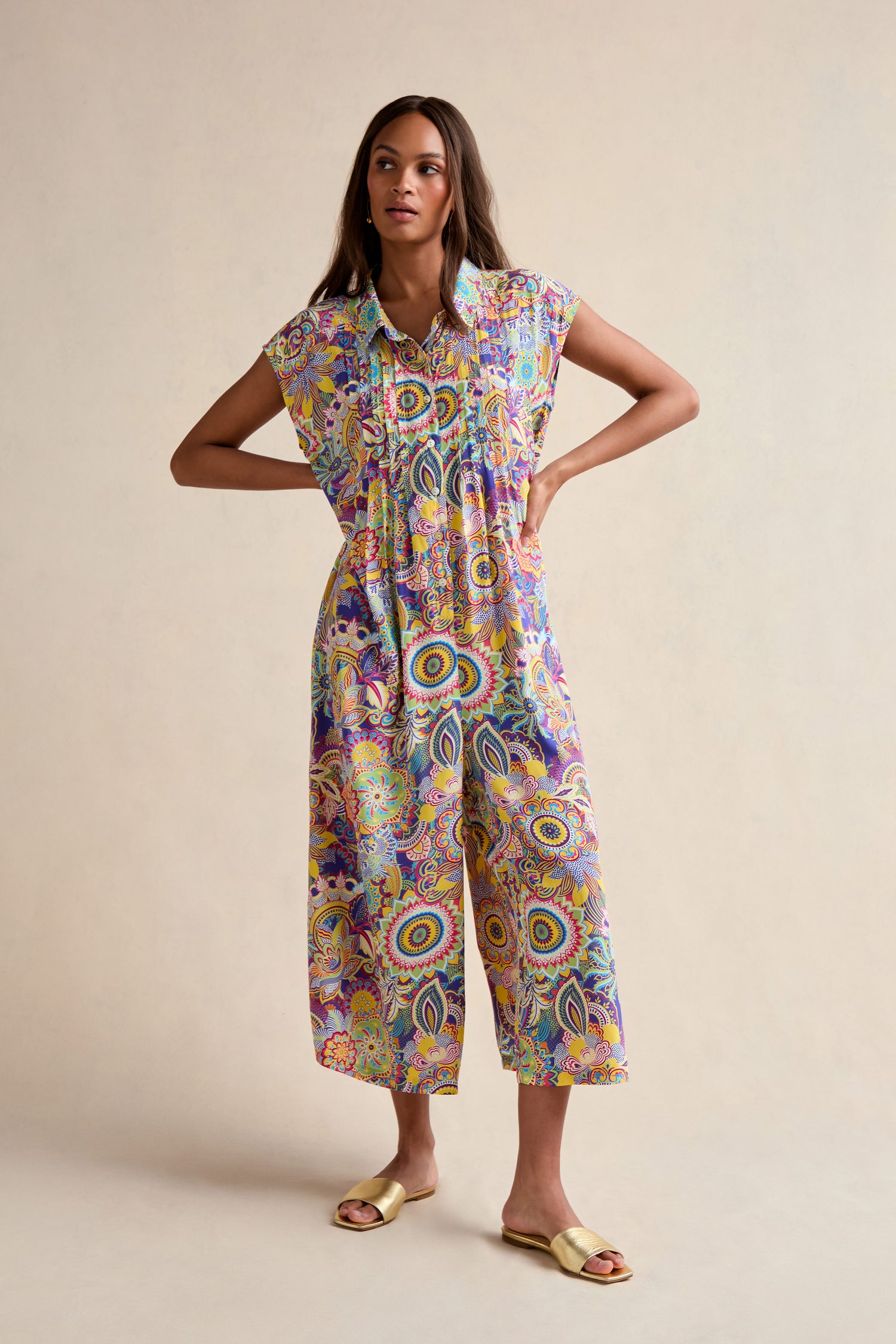 A woman stands with hands on hips, wearing the Summer Sarasa Jumpsuit—featuring wide legs and vibrant patterns—paired with yellow sandals against a plain beige background.