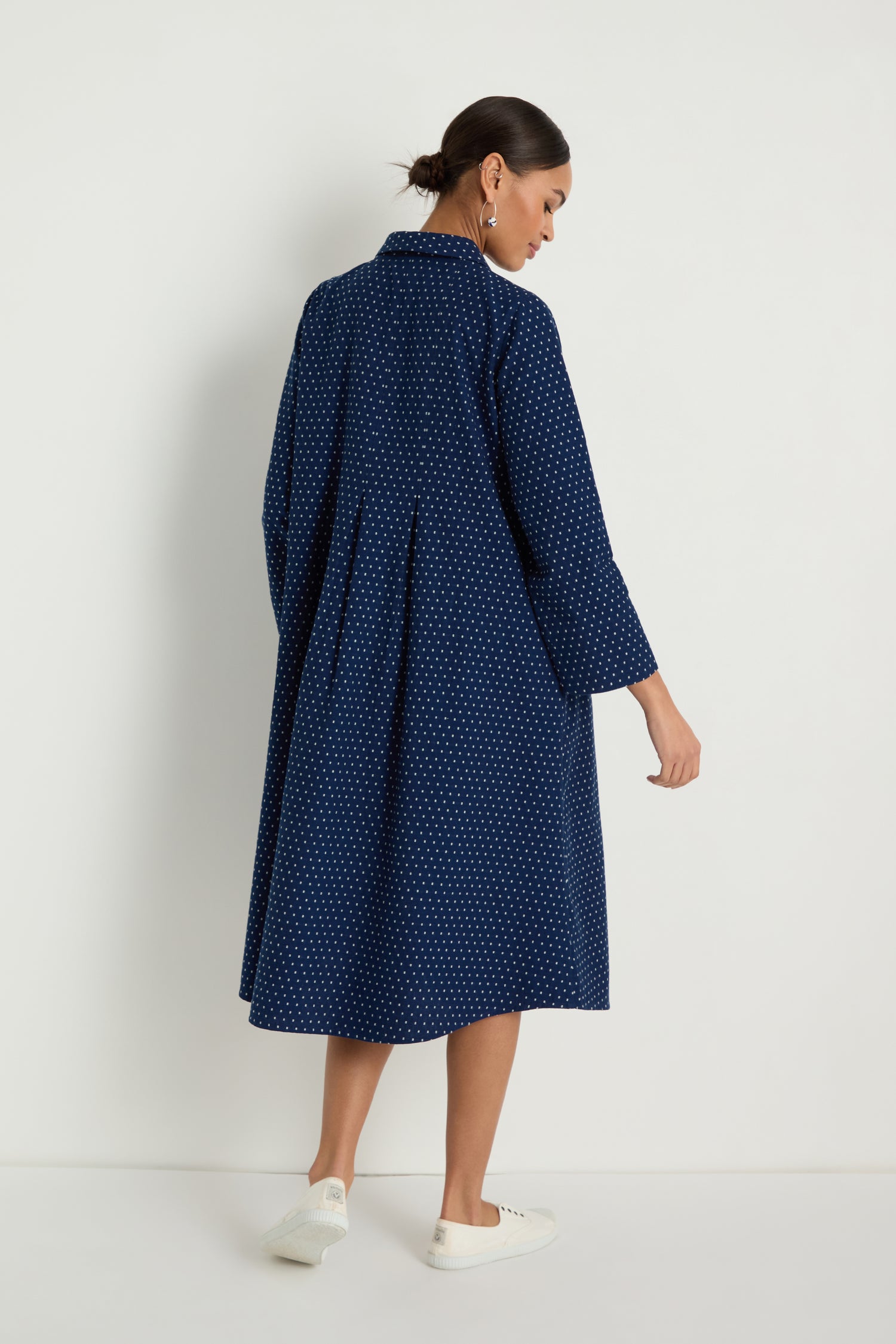 A woman faces away, wearing the Indigo Spot Cotton Shirt Dress—a navy knee-length, polka dot, long-sleeve cotton A-line with a relaxed fit. She pairs it with white shoes against a plain white background.