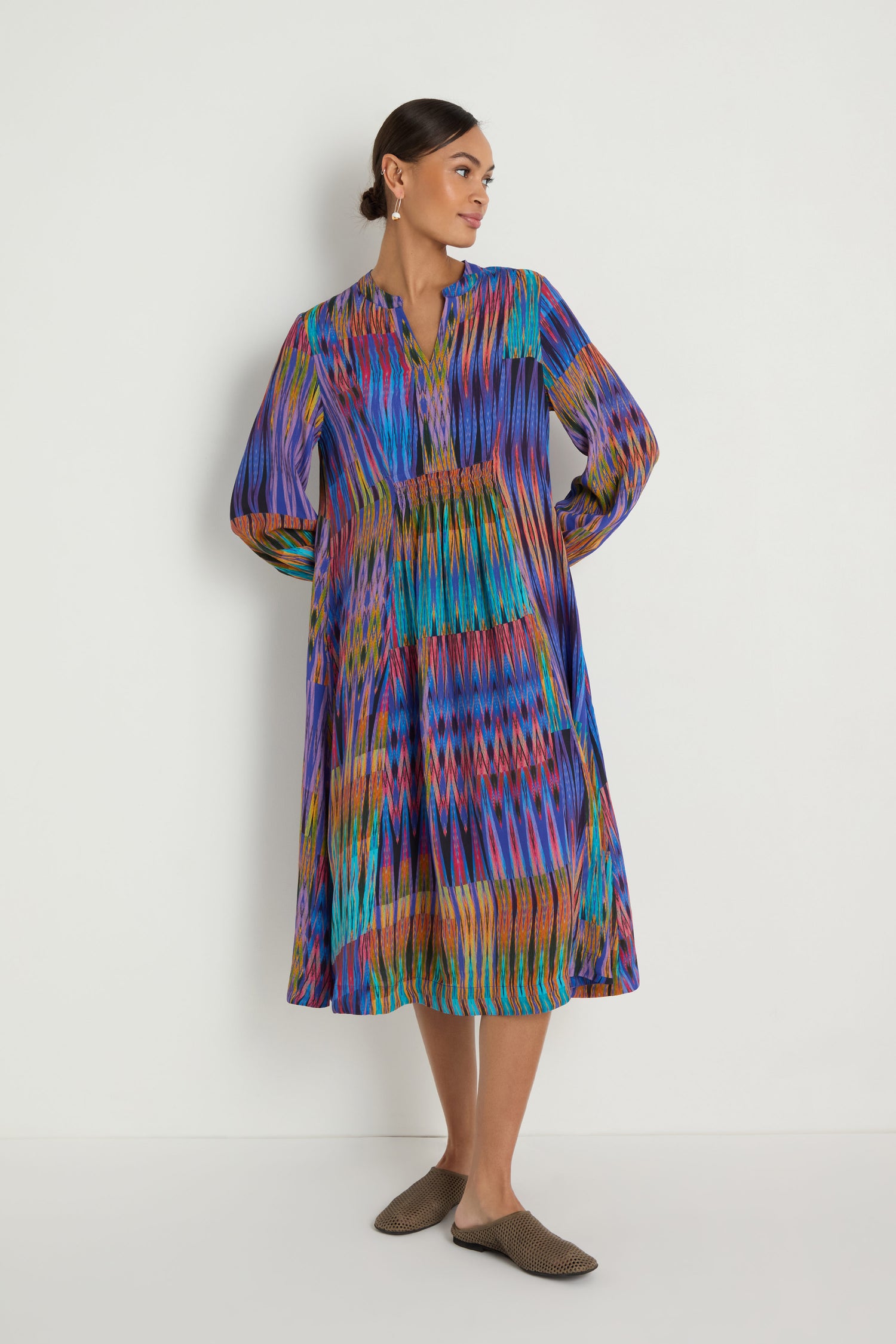 A woman models the Vivid Ikat Patchwork Dress, a long-sleeved, multicolored midi dress with a V-neck, standing against a plain white background and gazing to her left.