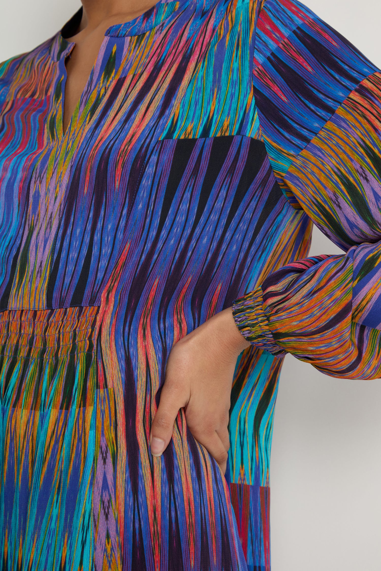 A person wearing the Vivid Ikat Patchwork Dress, a long-sleeved dress with a multicolored abstract linear pattern, rests their hand on their hip.