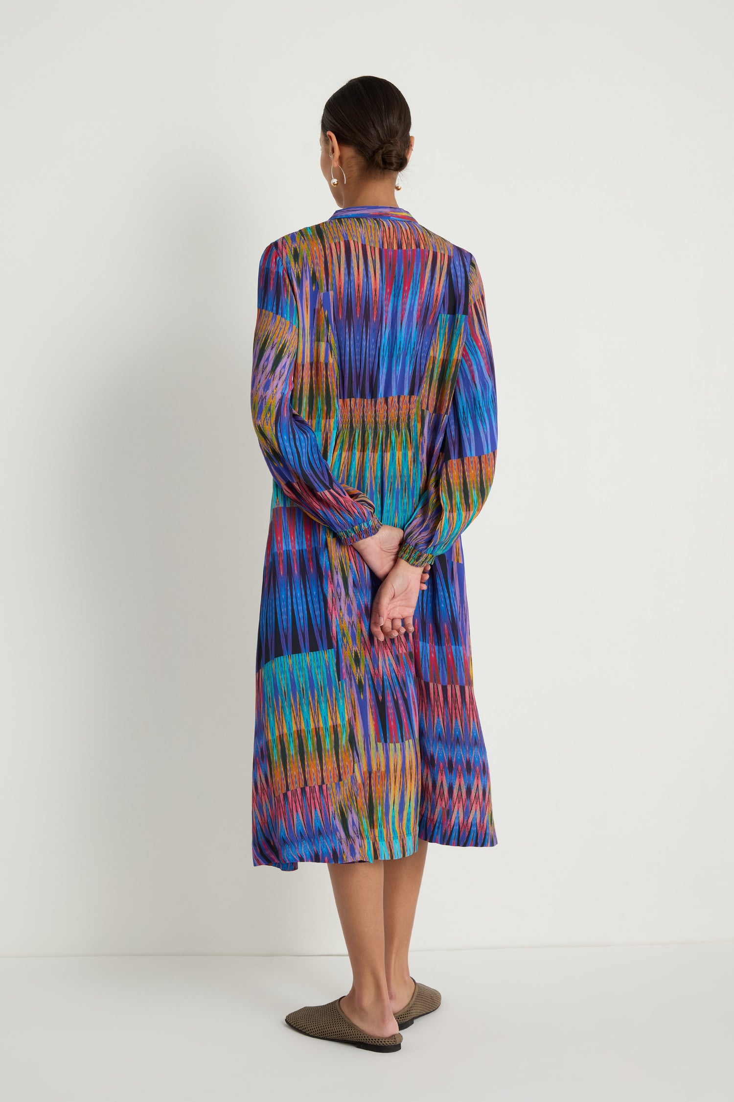 A woman stands facing a white wall with her hands behind her back, wearing the Vivid Ikat Patchwork Dress and brown flat shoes.