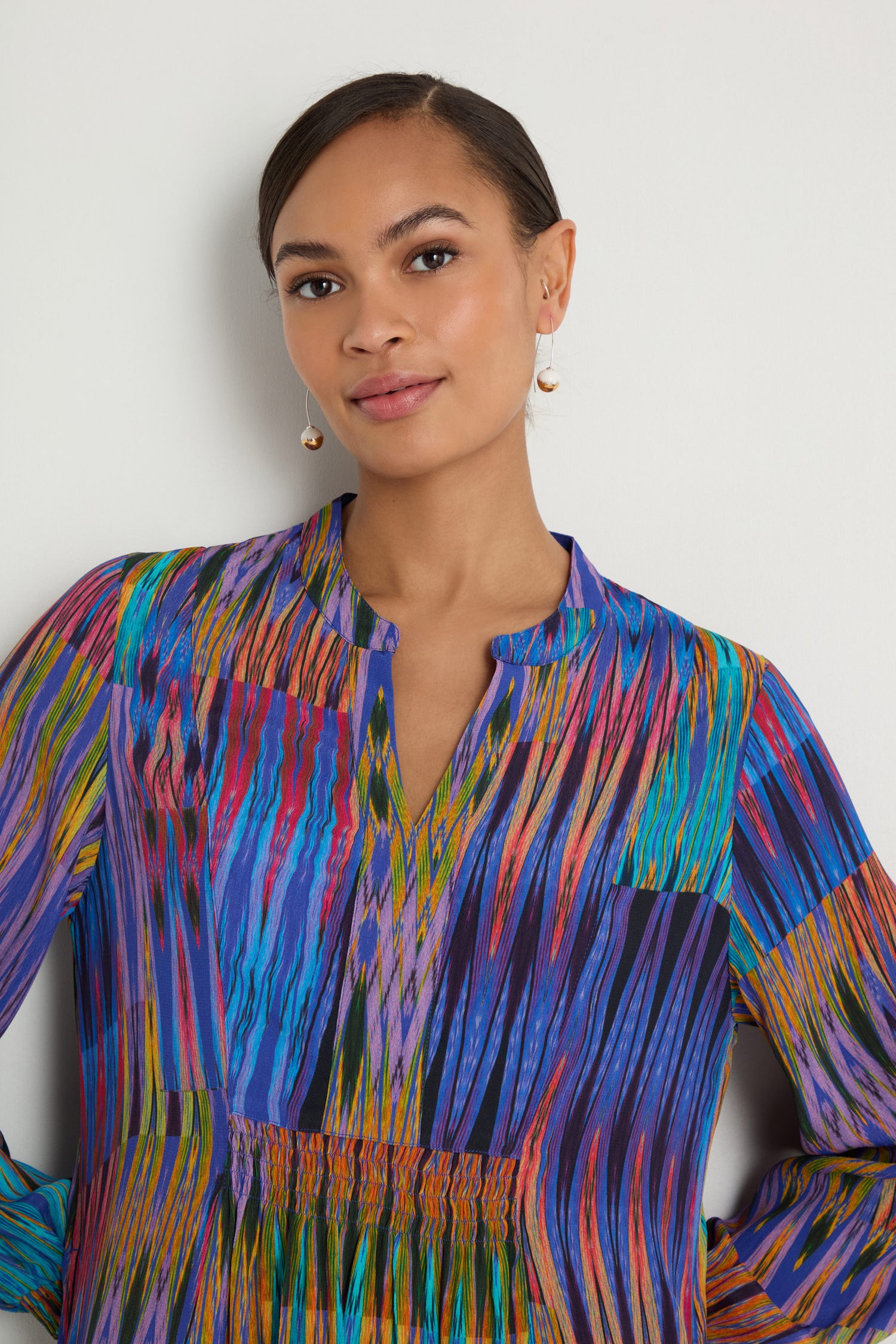 A woman with brown hair in a low bun wears the Vivid Ikat Patchwork Dress and drop earrings, standing against a white background.