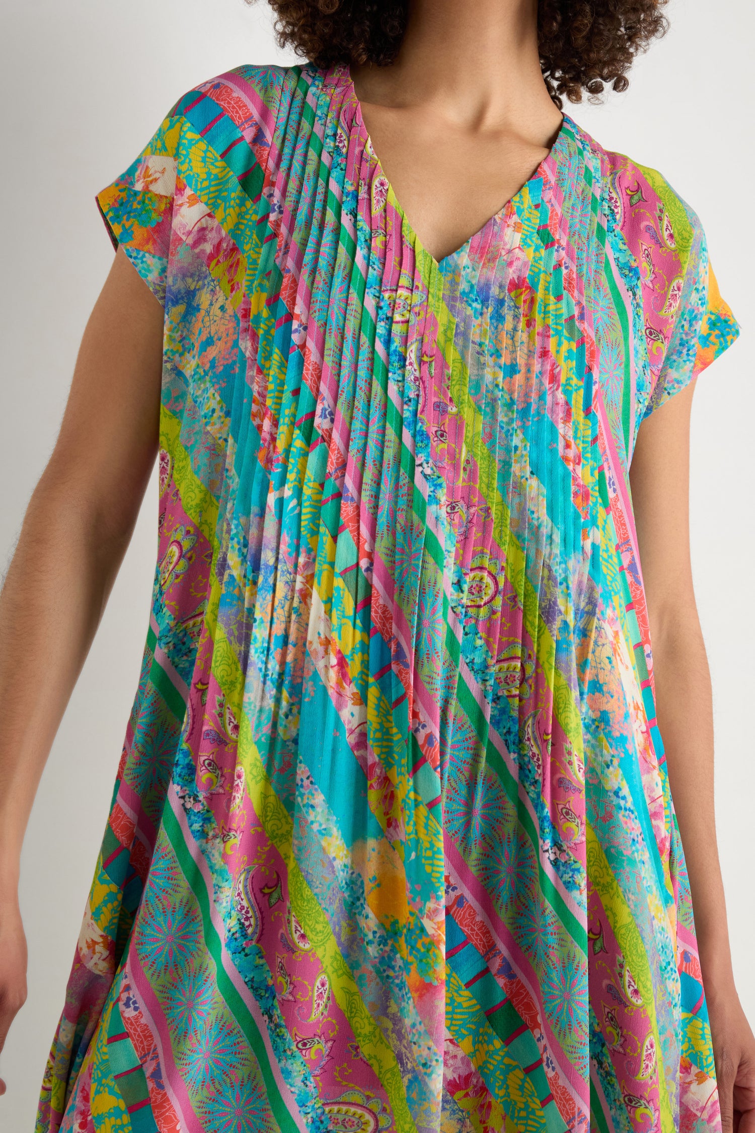 A person wears the Patchwork Stripe Pleated Dress, featuring colorful diagonal stripes and abstract patterns in pink, green, blue, and yellow with pleats, neckline detailing, and a flattering fit-and-flare silhouette.