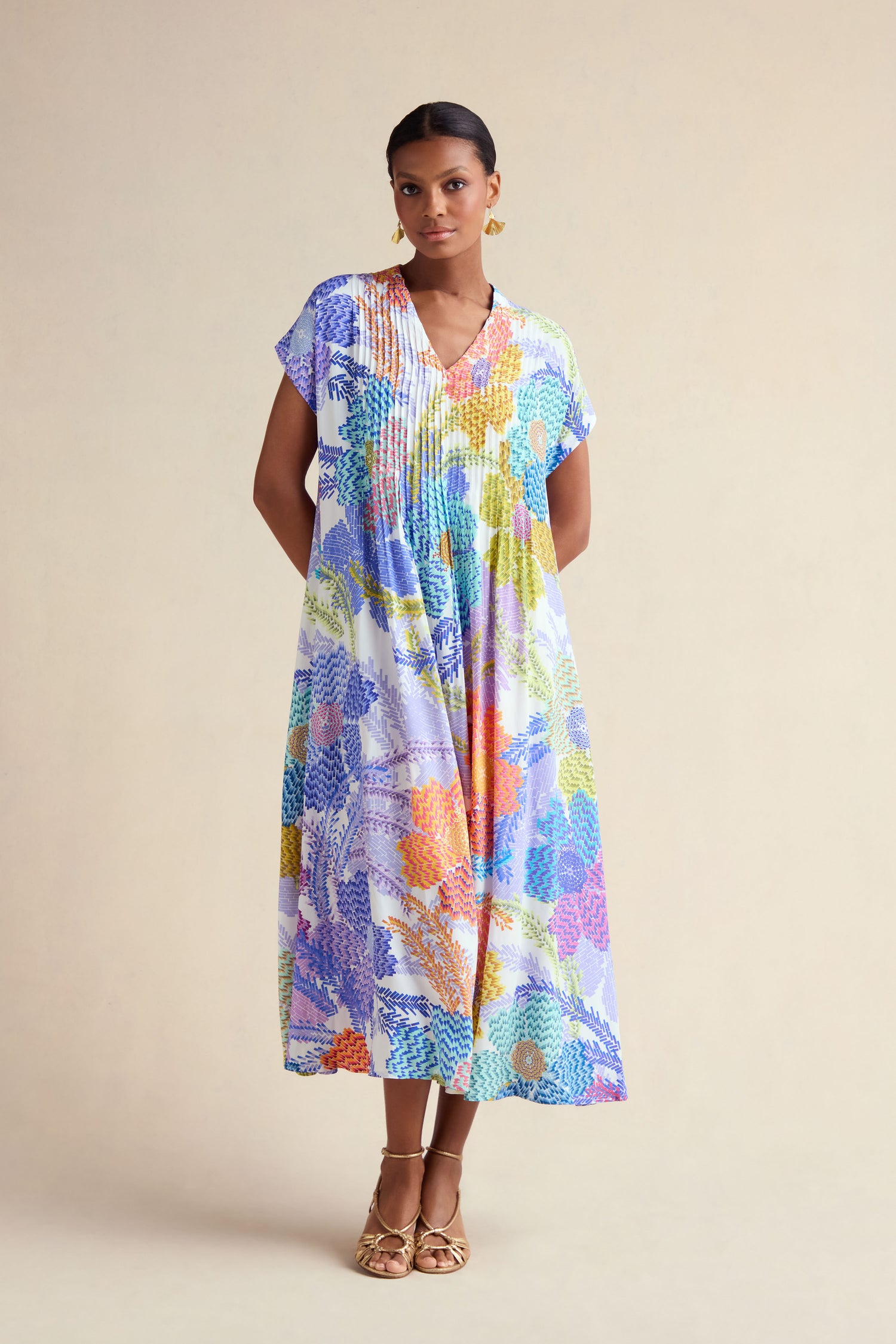 A woman poses against a beige background wearing the Pixelated Petals Print Dress with a loose fit, short sleeves, and gold sandals.