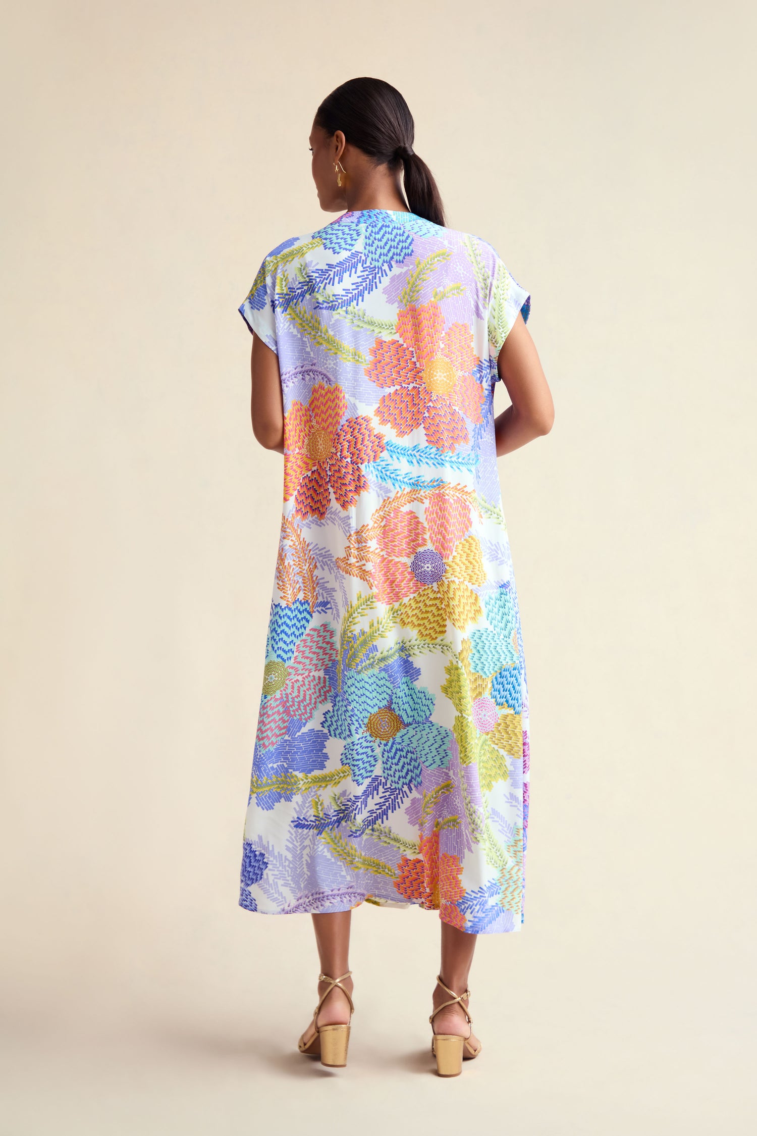 A woman with a ponytail and tan heeled sandals faces away, wearing the Pixelated Petals Print Dress—a loose, mid-length dress featuring a pixelated floral print—against a neutral background.