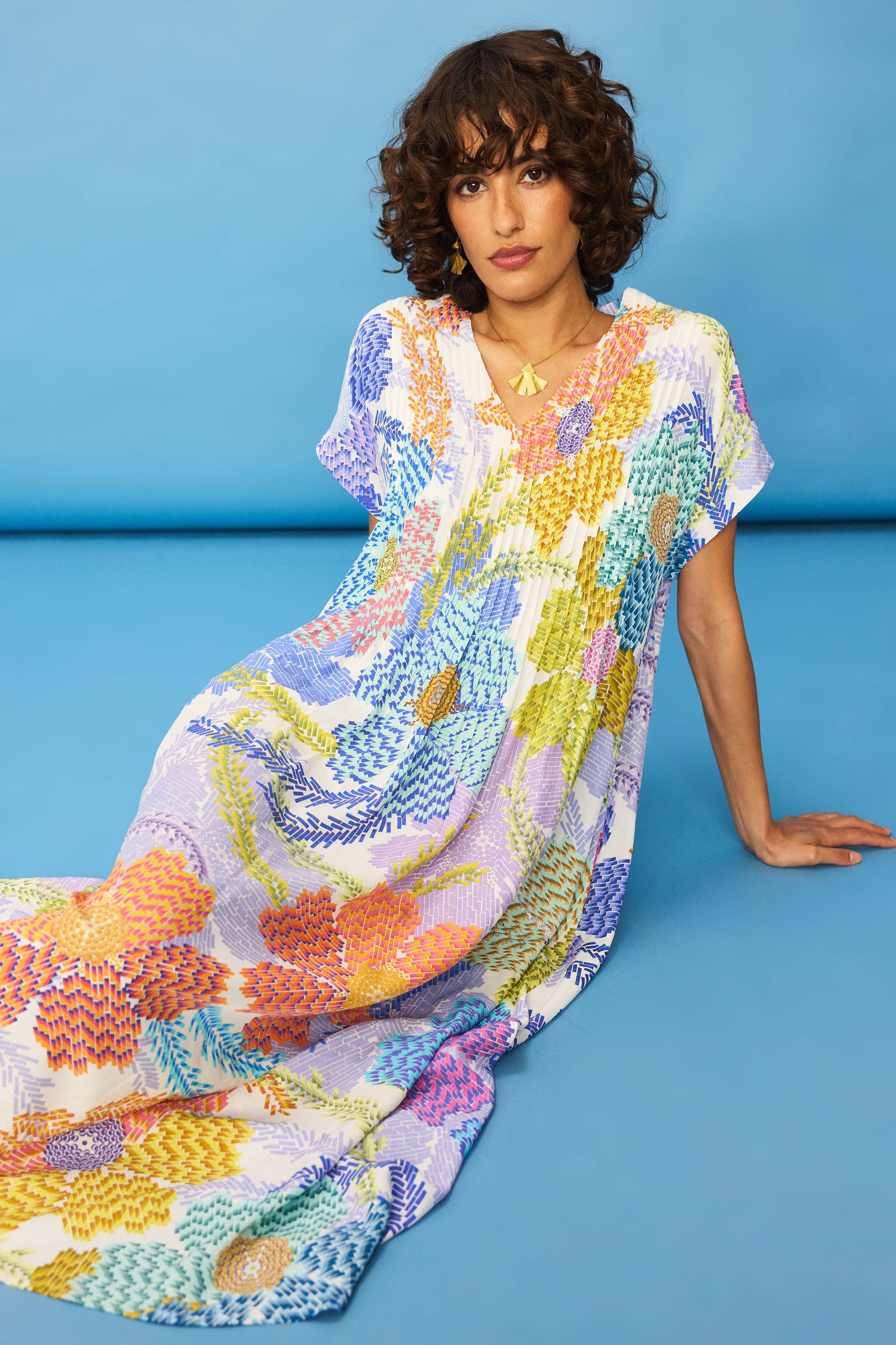 A person with curly hair sits on the floor against a blue background, wearing the loose-fitting Pixelated Petals Print Dress and a gold necklace.