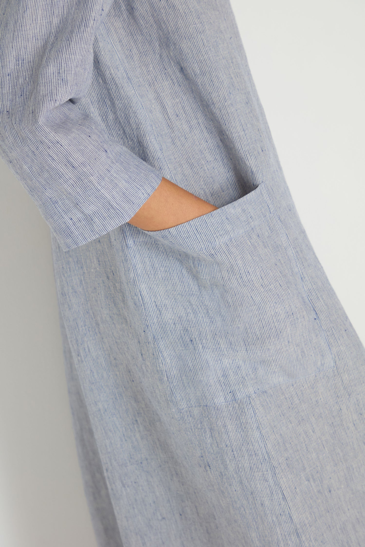 A side view of a hand in the pocket of the Ticking Linen Stripe Jumpsuit, featuring a light blue ticking stripe pattern and relaxed silhouette, set against a plain background.
