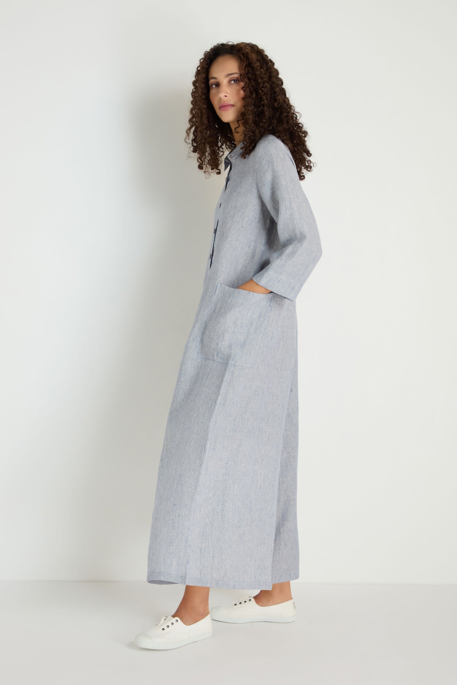 A woman with curly hair stands sideways, wearing the Ticking Linen Stripe Jumpsuit with pockets and white sneakers against a plain white background.