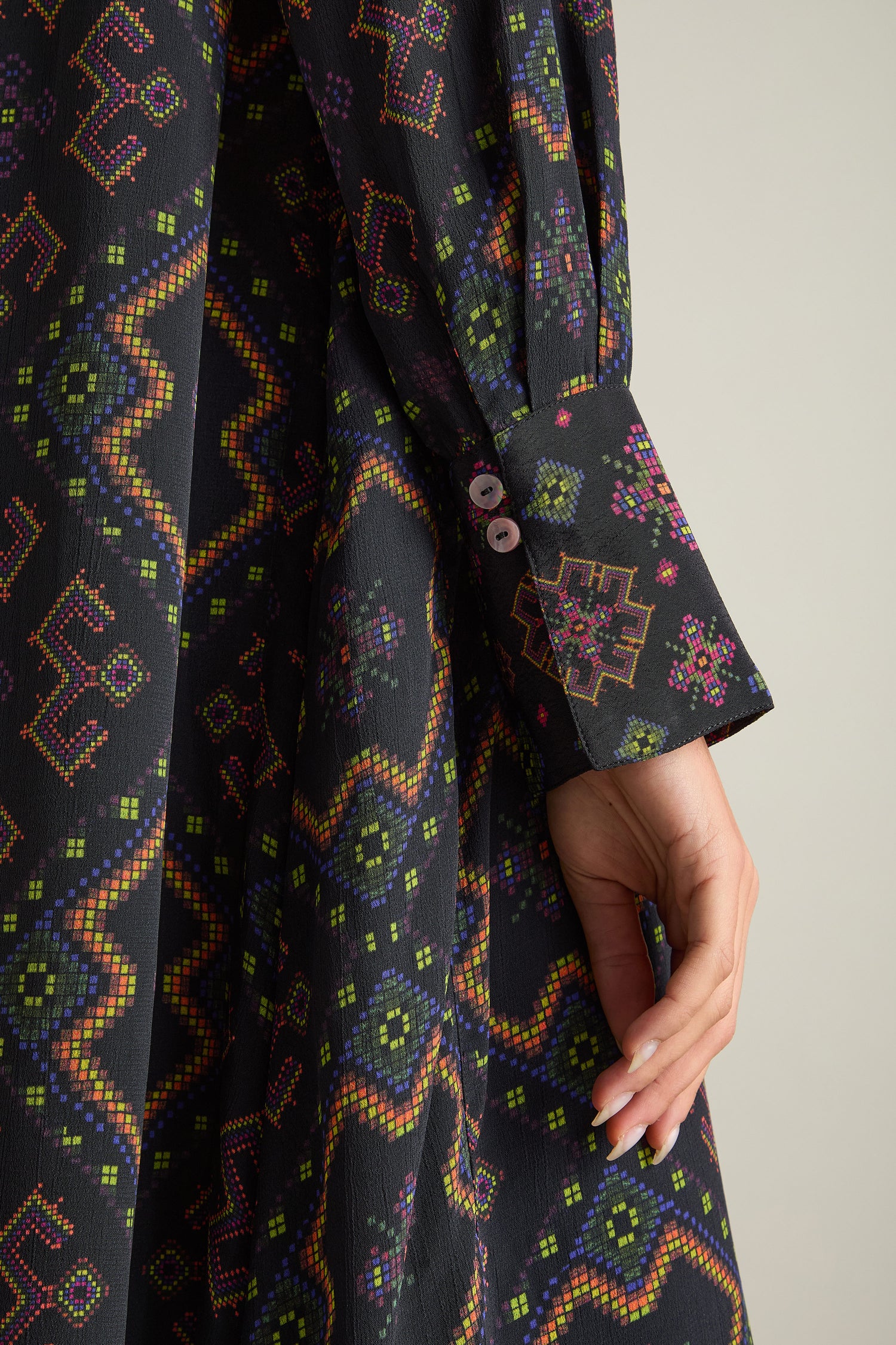 Close-up of a person's hand resting on the side of the silky crepe viscose Pixelated Jacquard Print Dress, adorned with intricate geometric and floral patterns in vibrant colors. This dress features long sleeves with buttoned cuffs.