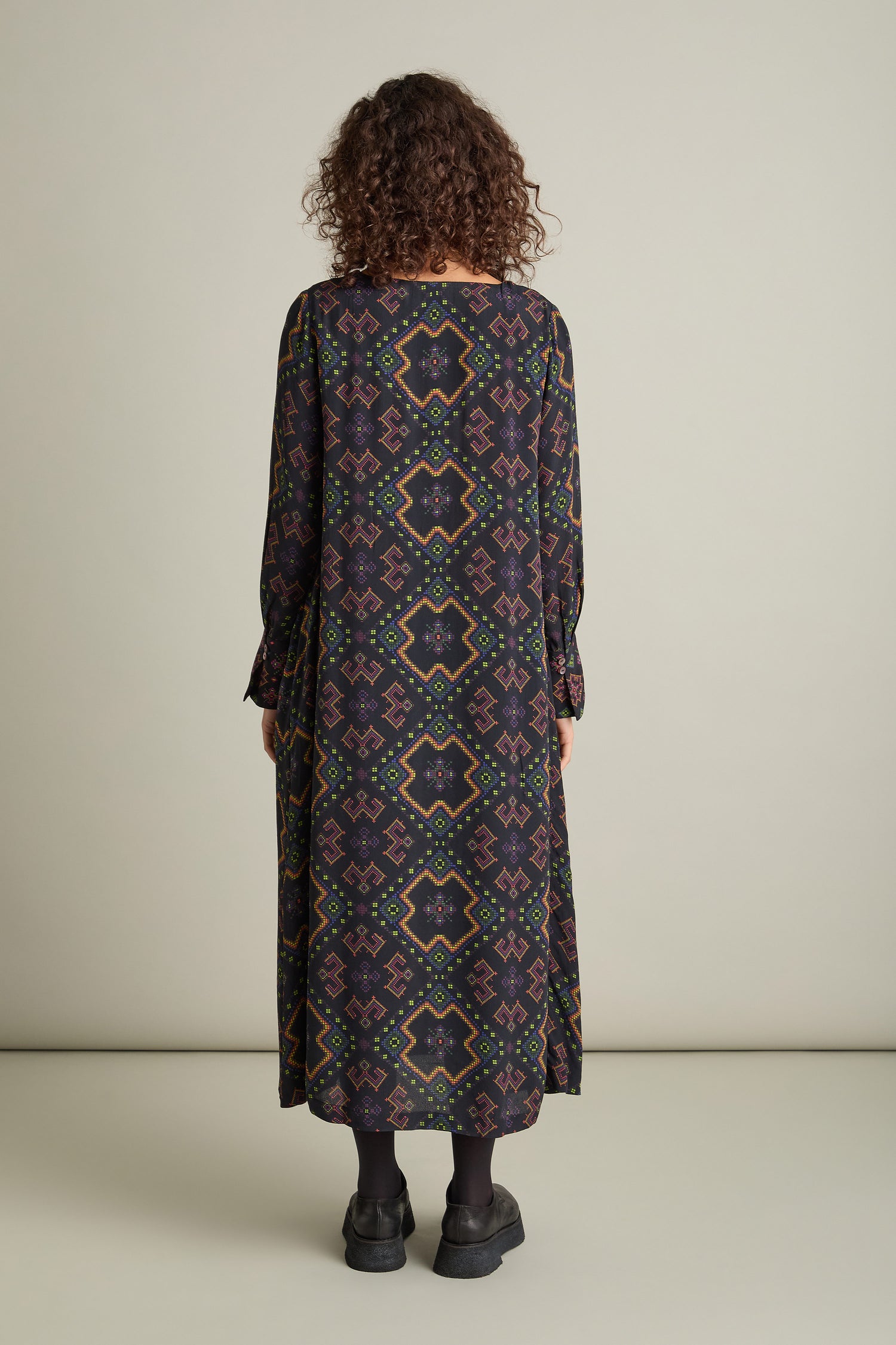 A person with curly hair stands facing away, donning a long Pixelated Jacquard Print Dress paired with dark shoes. The backdrop is plain and beige.