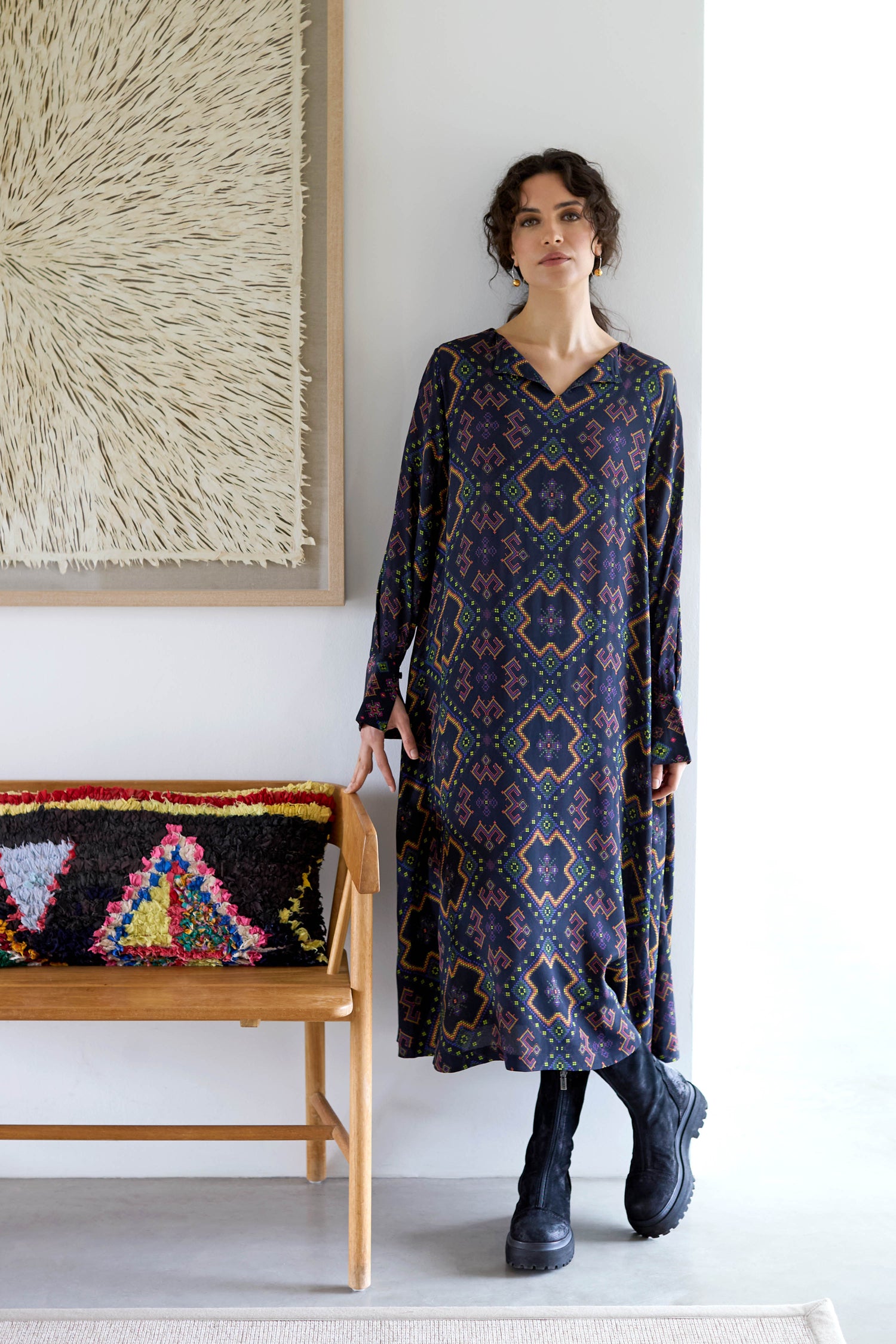 Wearing the Pixelated Jacquard Print Dress paired with boots, an individual stands next to a wooden bench adorned with colorful cushions, with abstract wall artwork serving as a backdrop.