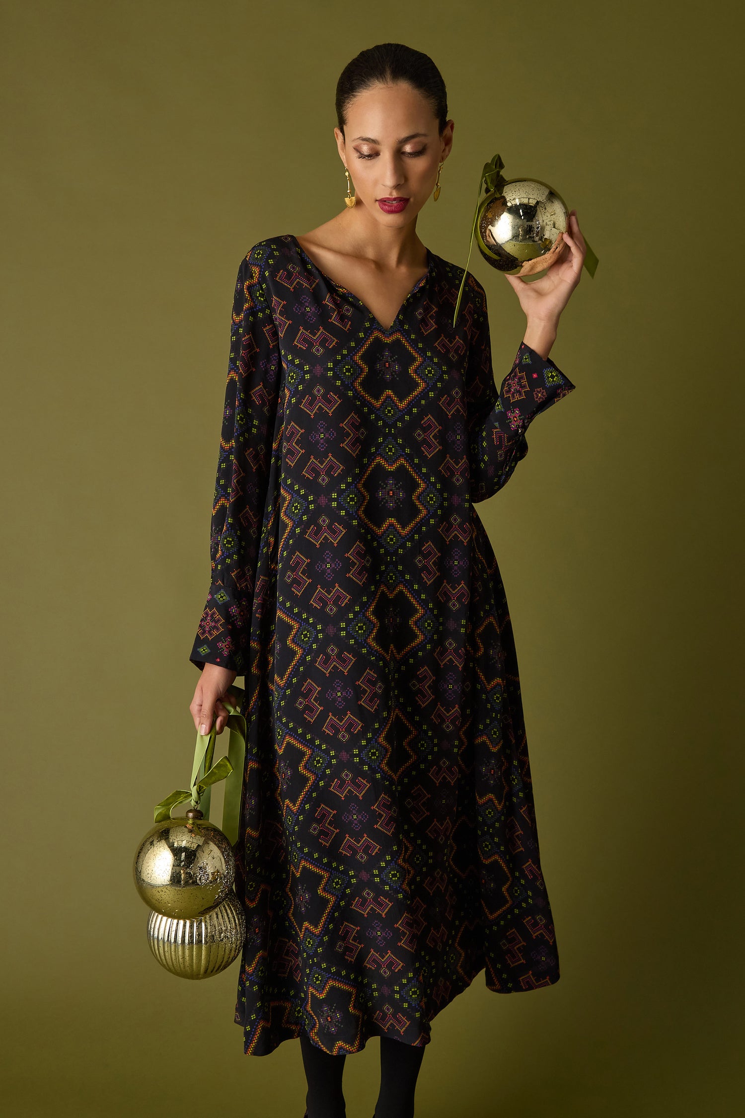 Dressed in a Pixelated Jacquard Print Dress featuring an A-line silhouette, an individual holds two ornaments in one hand and a single ornament in the other, set against a green background.