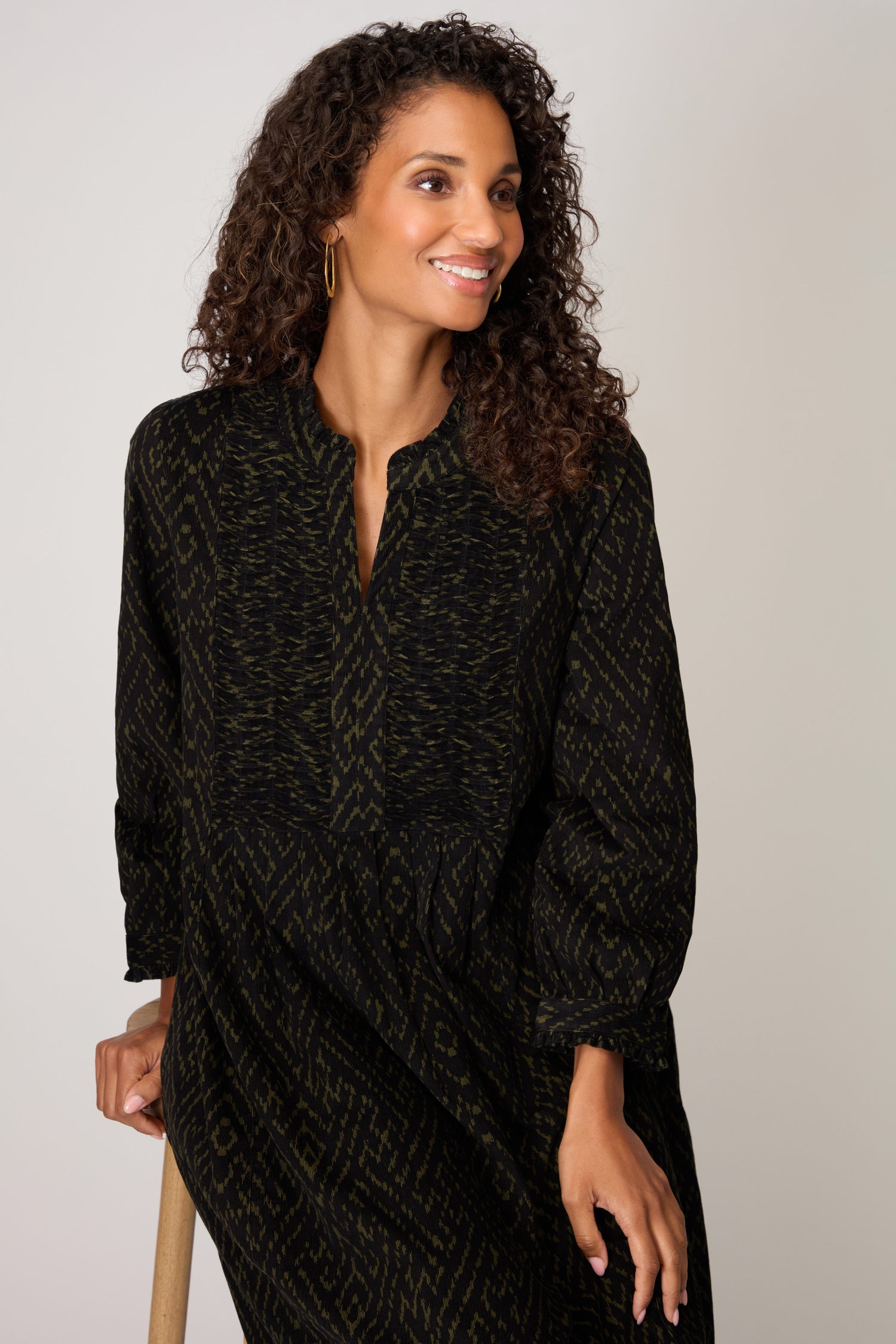 A woman with curly hair wears the Ornate Textured Black and Khaki Cord Dress, featuring a relaxed silhouette and long sleeves, paired with gold hoop earrings, standing against a plain light background.