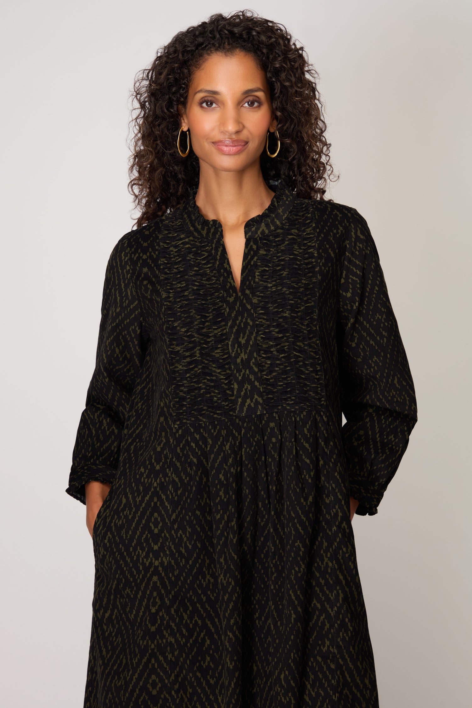 A woman with curly hair poses facing the camera with her hands in her pockets, wearing the Ornate Textured Black and Khaki Cord Dress in a relaxed silhouette, against a plain backdrop.