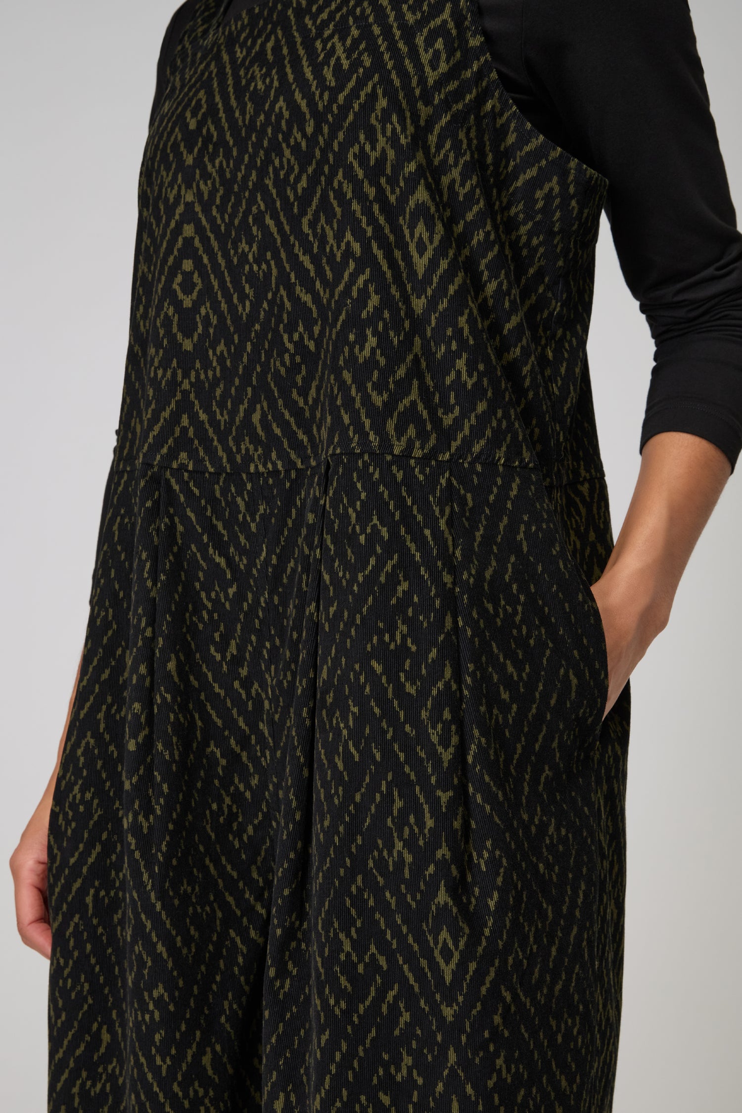 A person wears the Ornate Textured Cord Jumpsuit—sleeveless and patterned in black and dark green with side pockets—over a black long-sleeve top, creating an effortless statement with relaxed tailoring.