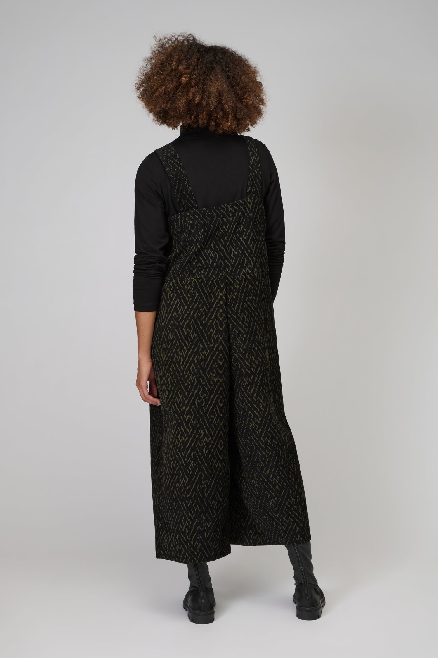 Person facing away, wearing a statement piece: the Ornate Textured Cord Jumpsuit over a black turtleneck and boots, standing against a plain light background.