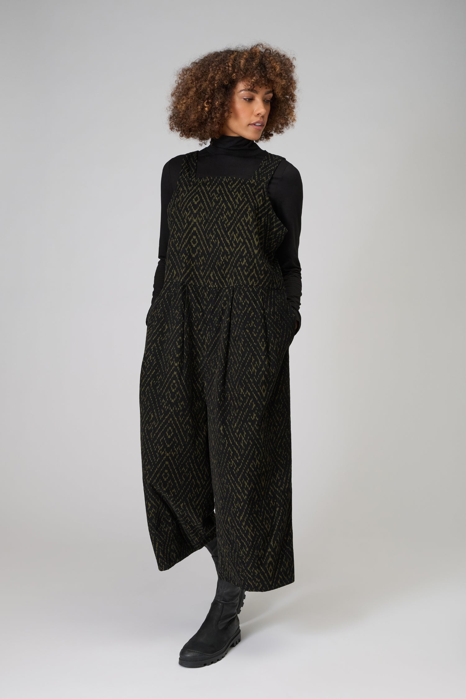 A person with curly hair wears a black turtleneck and the Ornate Textured Cord Jumpsuit with pockets, styled with black boots, standing against a plain light background.