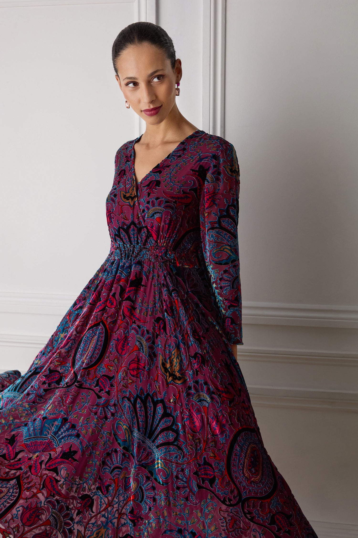 A woman wearing the Winter Paisley Devore Flared Dress, characterized by its long and colorful design, stands against a white-paneled wall. The A-line silhouette of the dress complements her form as she gazes to the side with a slight smile.