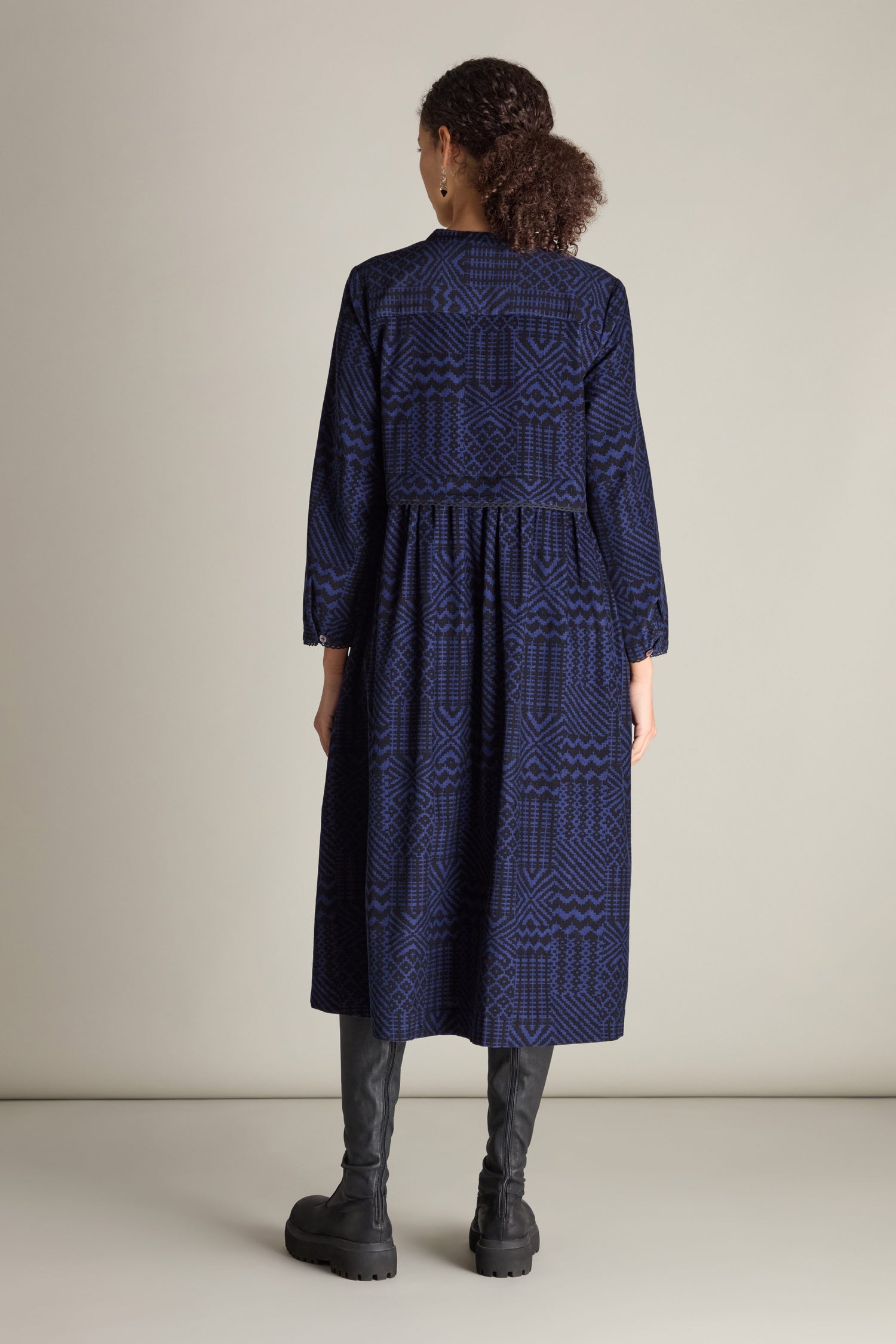 A person with curly hair stands facing away, wearing a long-sleeved Mono Ikat Printed Cord Dress in dark blue paired with black knee-high boots against a plain background.