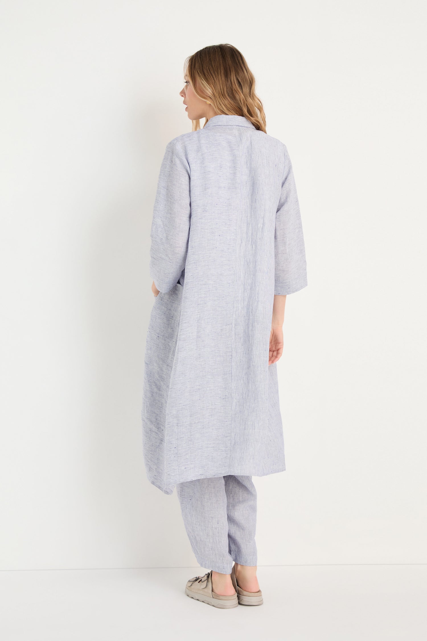A person stands facing away, wearing a relaxed Ticking Linen Stripe Shirt Dress in light gray with matching pants and slip-on shoes against a plain white background.