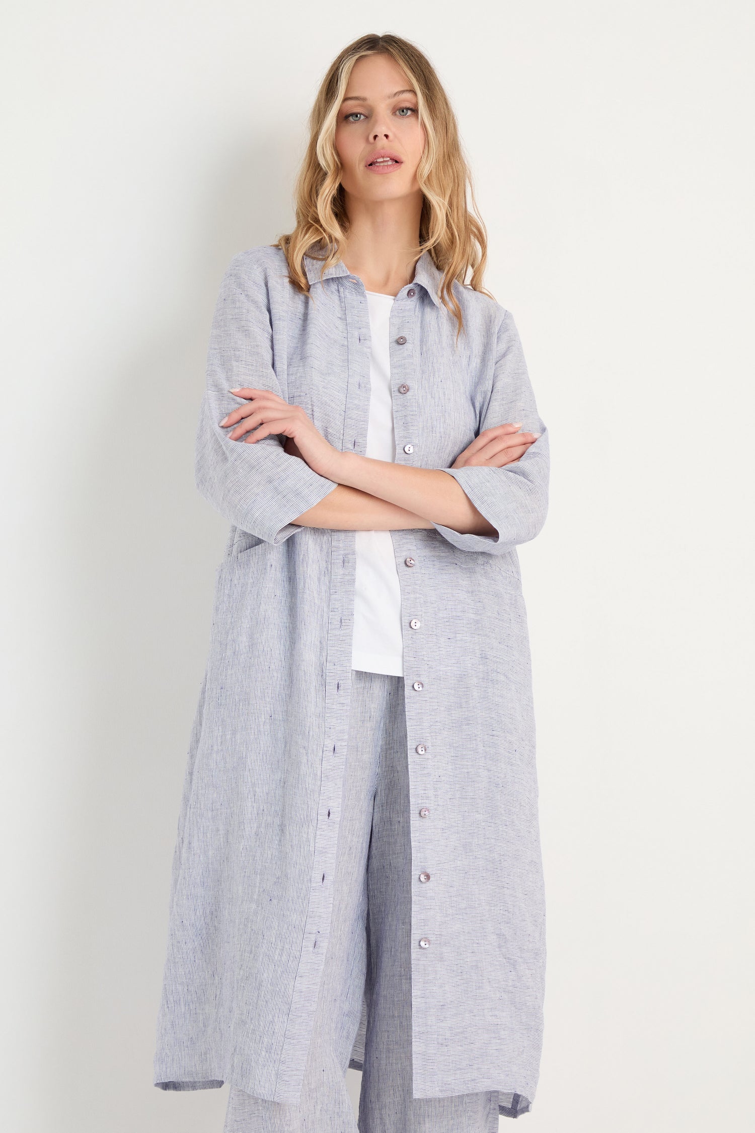 A woman stands against a white wall with her arms crossed, wearing the Ticking Linen Stripe Shirt Dress over a white top and matching wide-leg pants.