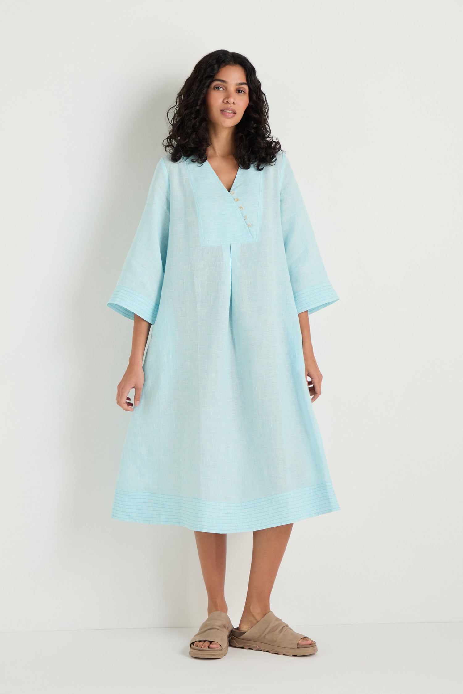 A woman stands against a plain background wearing the Ticking Stripe Linen Button Placket Dress, which has an A-line silhouette and light blue stripes, paired with beige slide sandals.