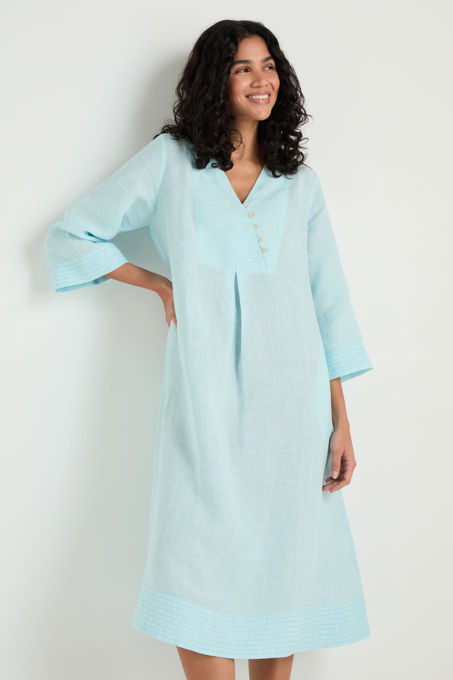 A woman stands against a plain white wall wearing the Ticking Stripe Linen Button Placket Dress, a light blue knee-length style with three-quarter sleeves and front button placket.