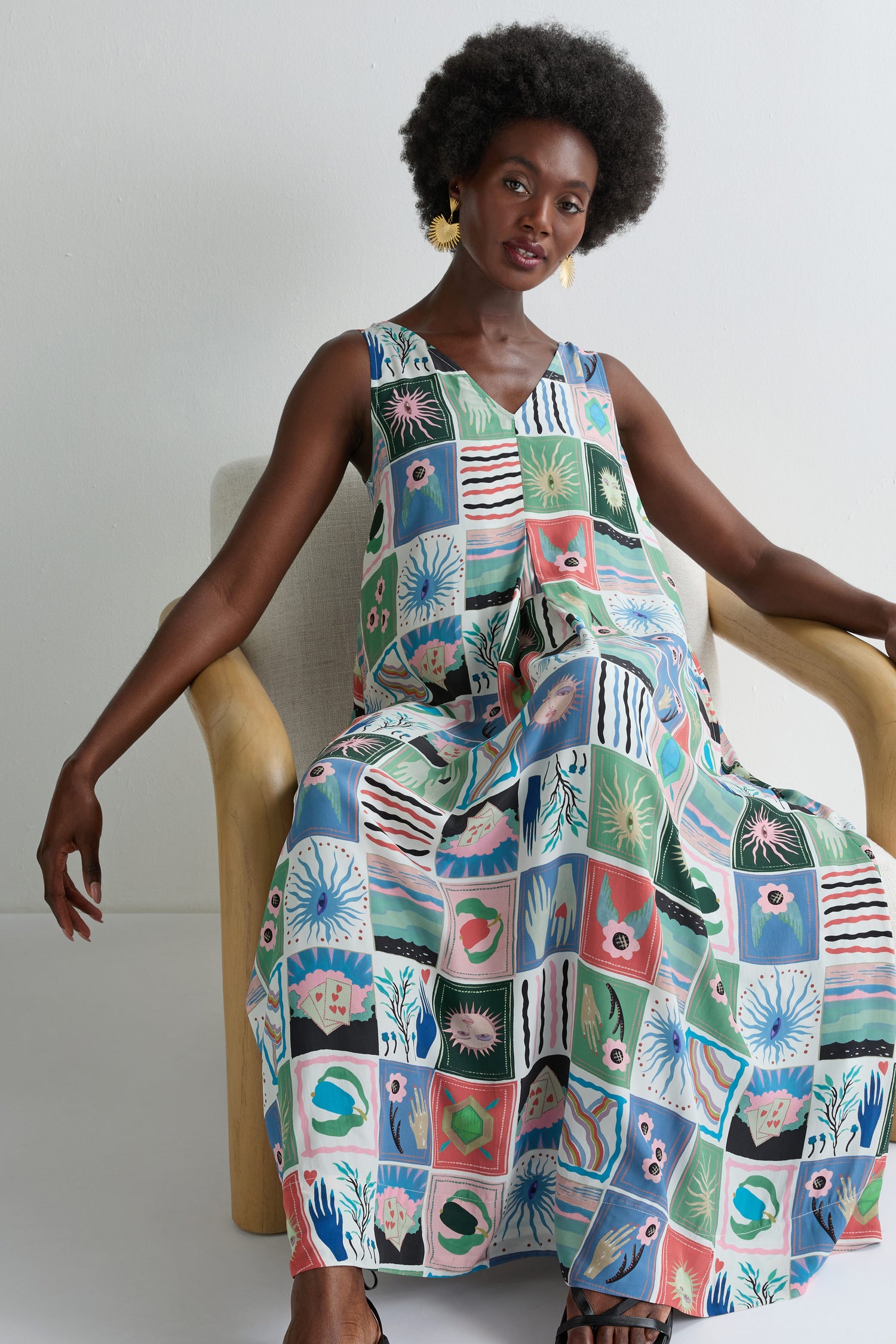 A woman wearing the Mystical Collage Sleeveless Dress with a vibrant square pattern sits on a light chair in front of a plain white wall.
