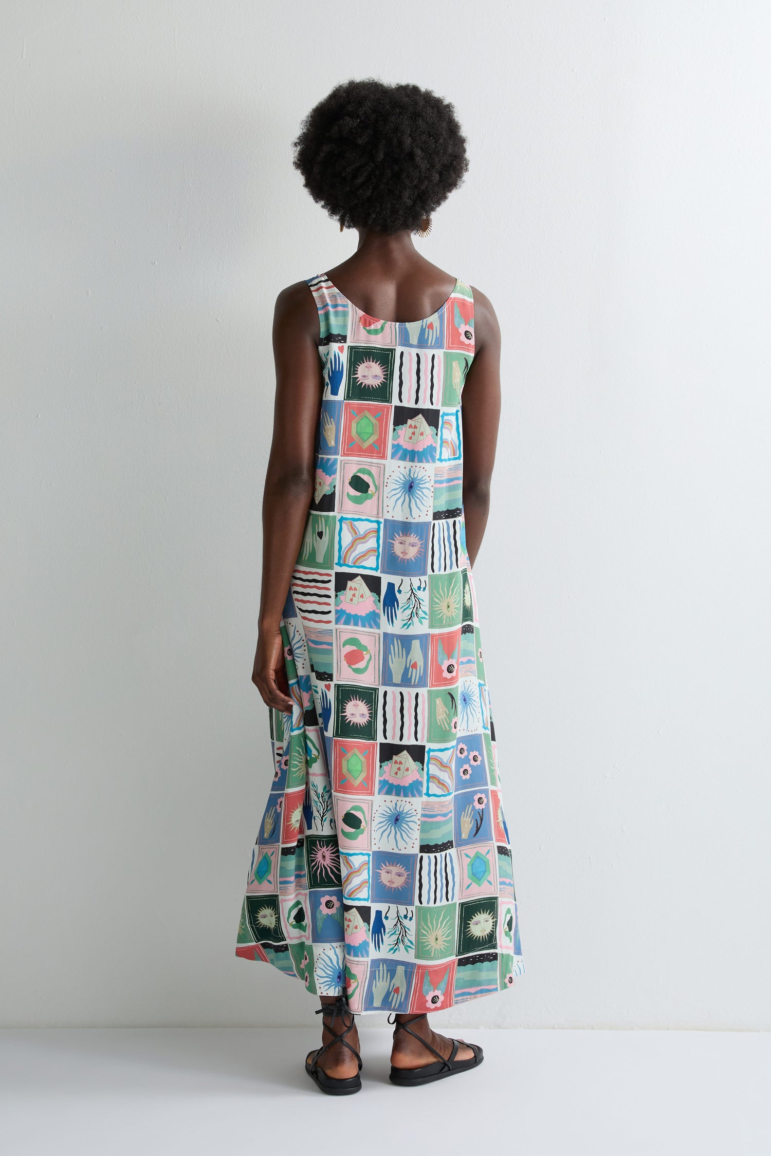 Someone stands facing a white wall, wearing the Mystical Collage Sleeveless Dress featuring a vibrant geometric and botanical pattern.