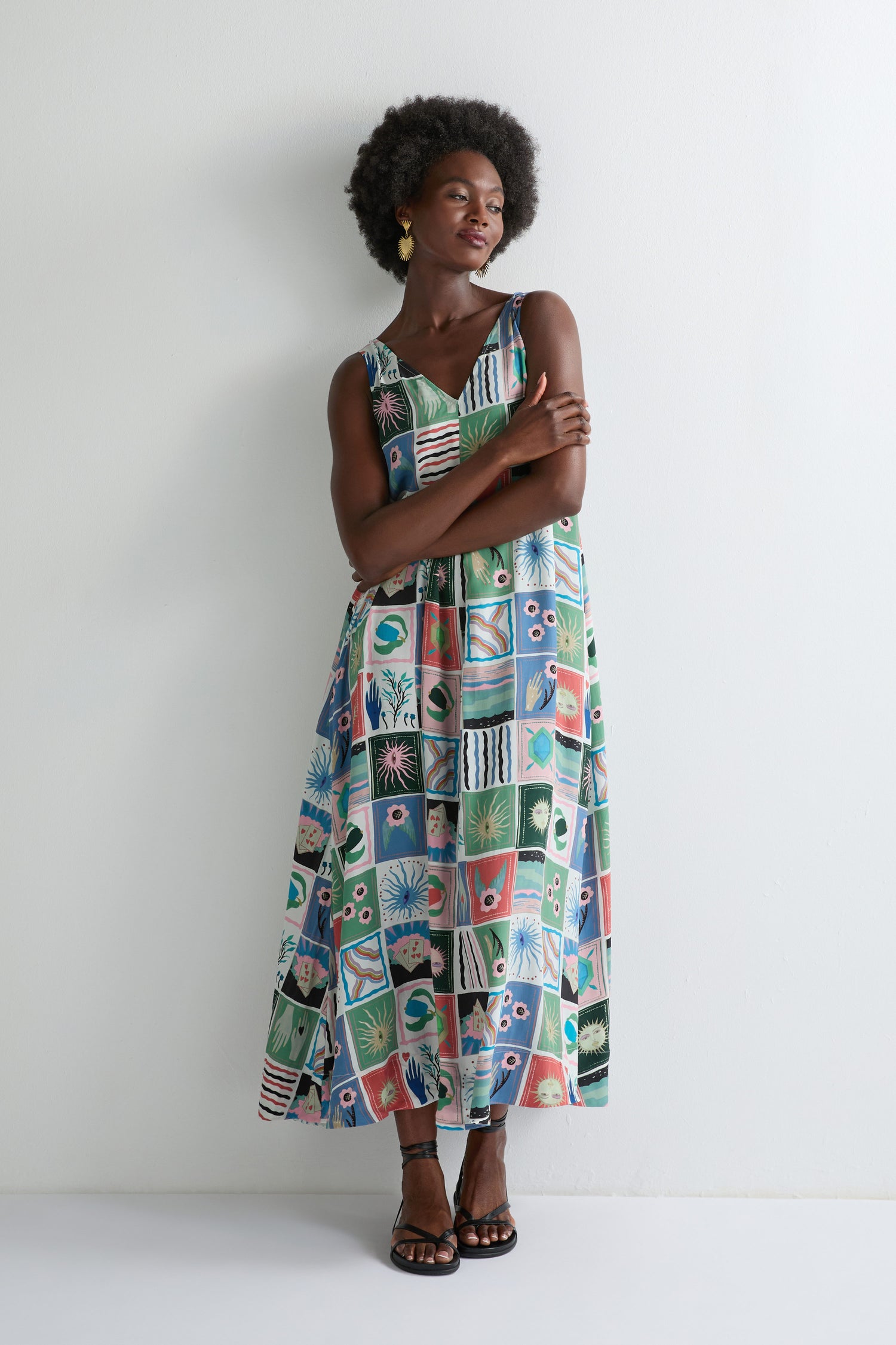 A woman stands against a plain white wall with her arms crossed, wearing the Mystical Collage Sleeveless Dress featuring vibrant geometric and floral patchwork patterns—perfect for summer.