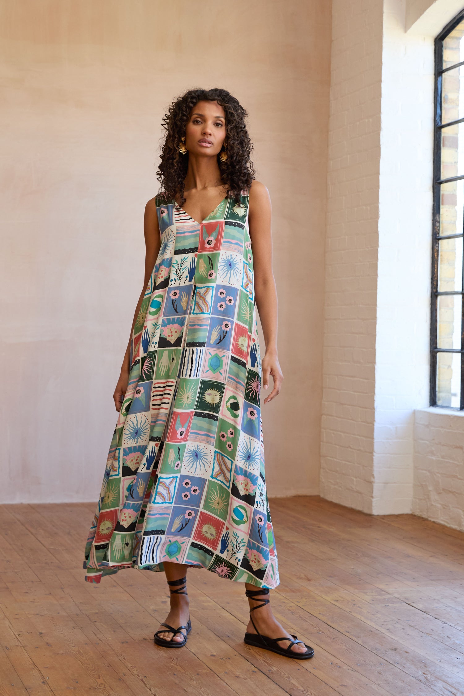 Indoors on wooden floors and against white brick walls, a woman wears the Mystical Collage Sleeveless Dress—a loose, A-line dress with a colorful patchwork pattern—paired with black sandals.