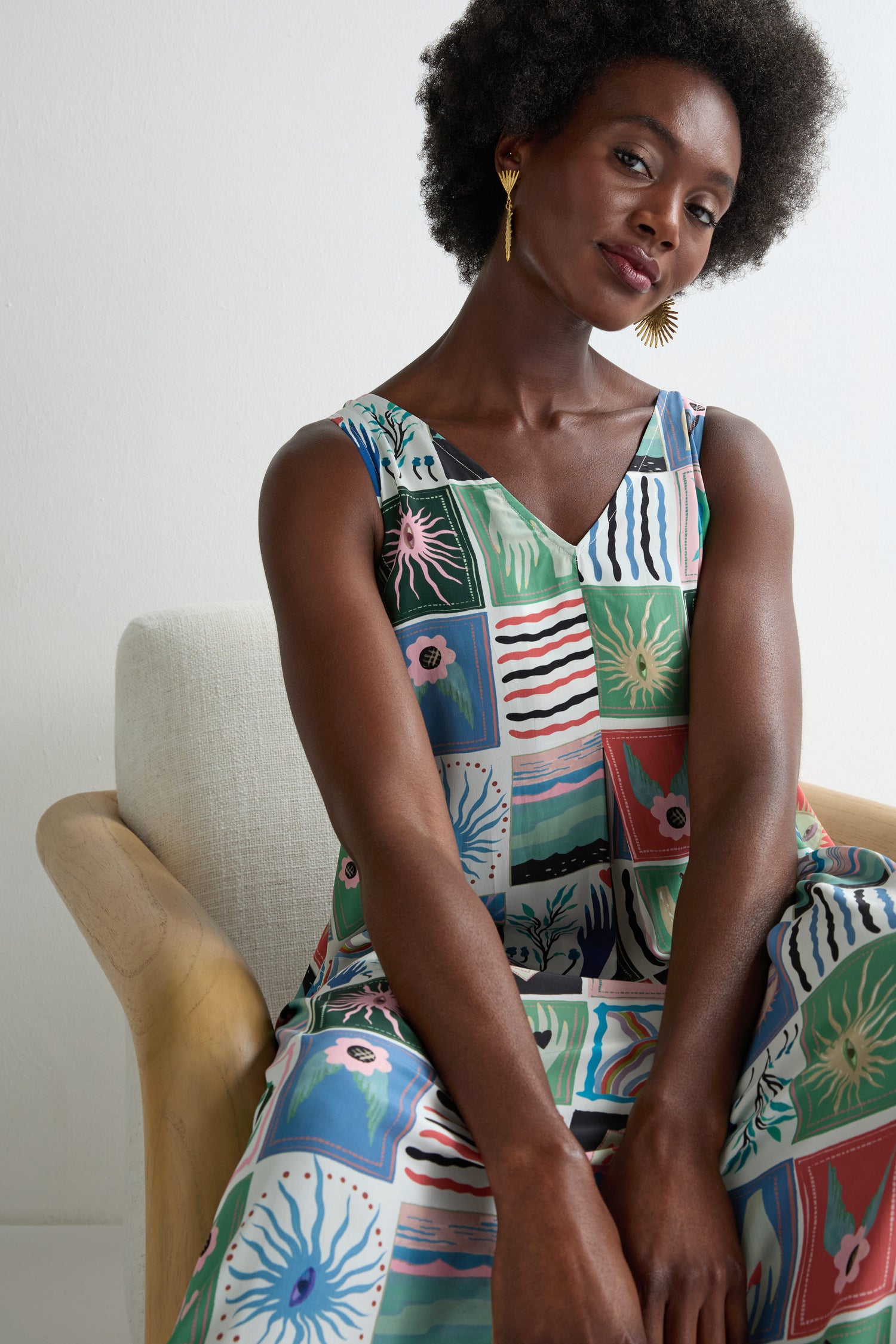 A woman with short curly hair sits on a light chair, wearing the Mystical Collage Sleeveless Dress—a colorful, patterned summer dress—paired with gold statement earrings.