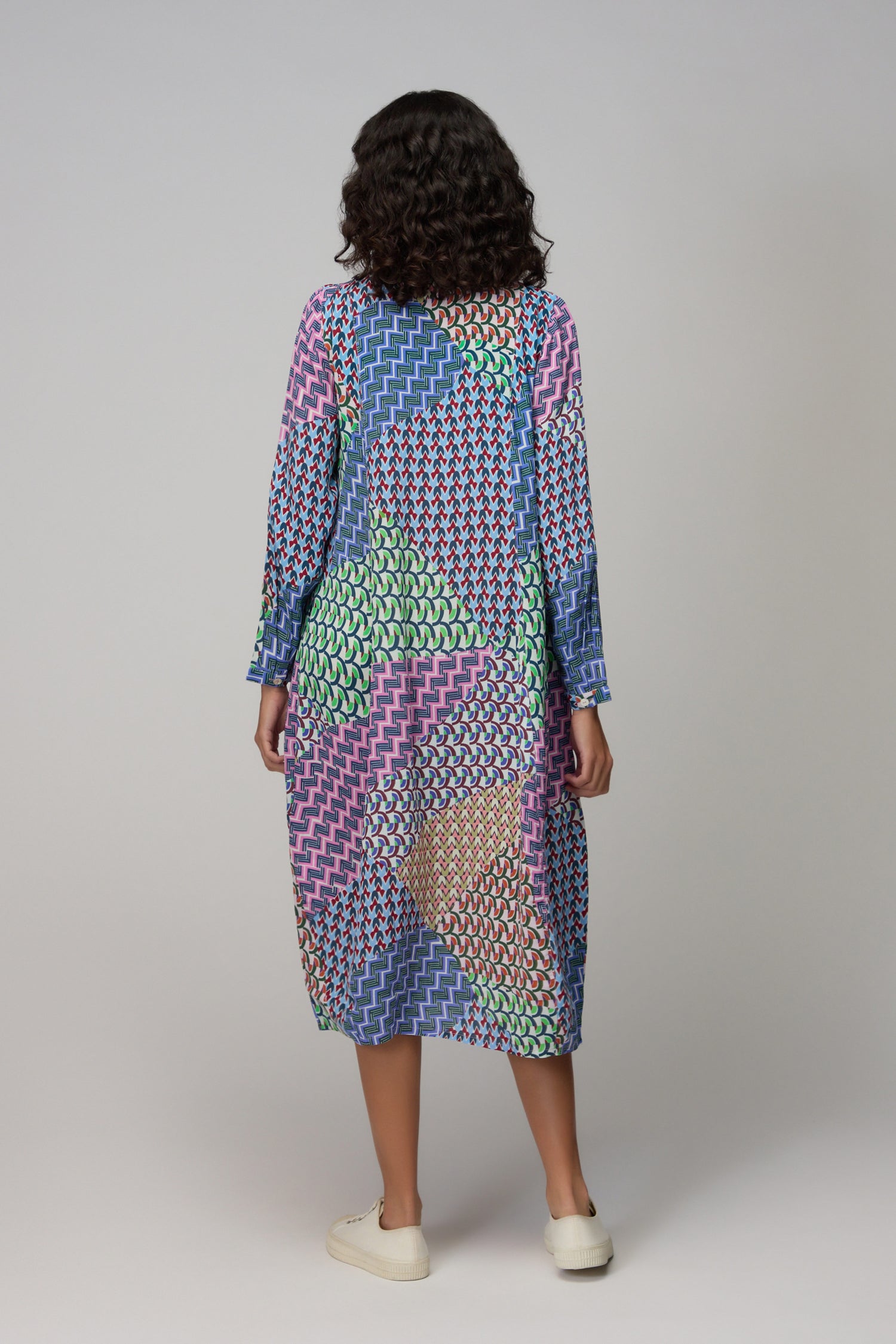 A woman poses against a plain background in the Geo Patchwork Print Shirt Dress, a statement piece with long sleeves and a button-up front, styled with white sneakers for a fresh modern look.