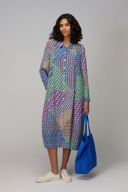 A woman stands against a plain background wearing the Geo Patchwork Print Shirt Dress, featuring colorful geometric patterns and long sleeves, paired with white sneakers.