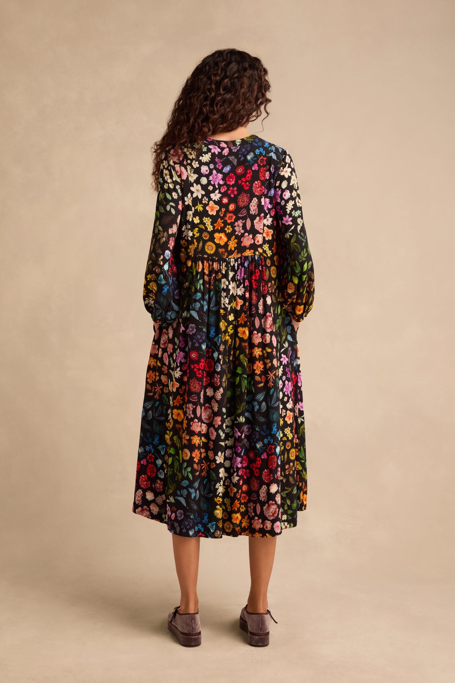 A woman with curly hair stands facing away, wearing the Midnight Garden Viscose Dress—an ankle-length, long-sleeved floral dress with a colorful pattern—and gray shoes against a beige background.