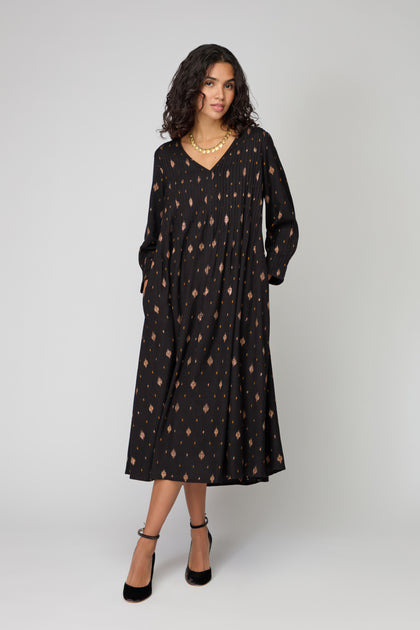 A woman in the Diamond Lurex Pleated Dress—black and gold with a subtle geometric print—paired with black heels and a gold necklace, stands against a plain light background.