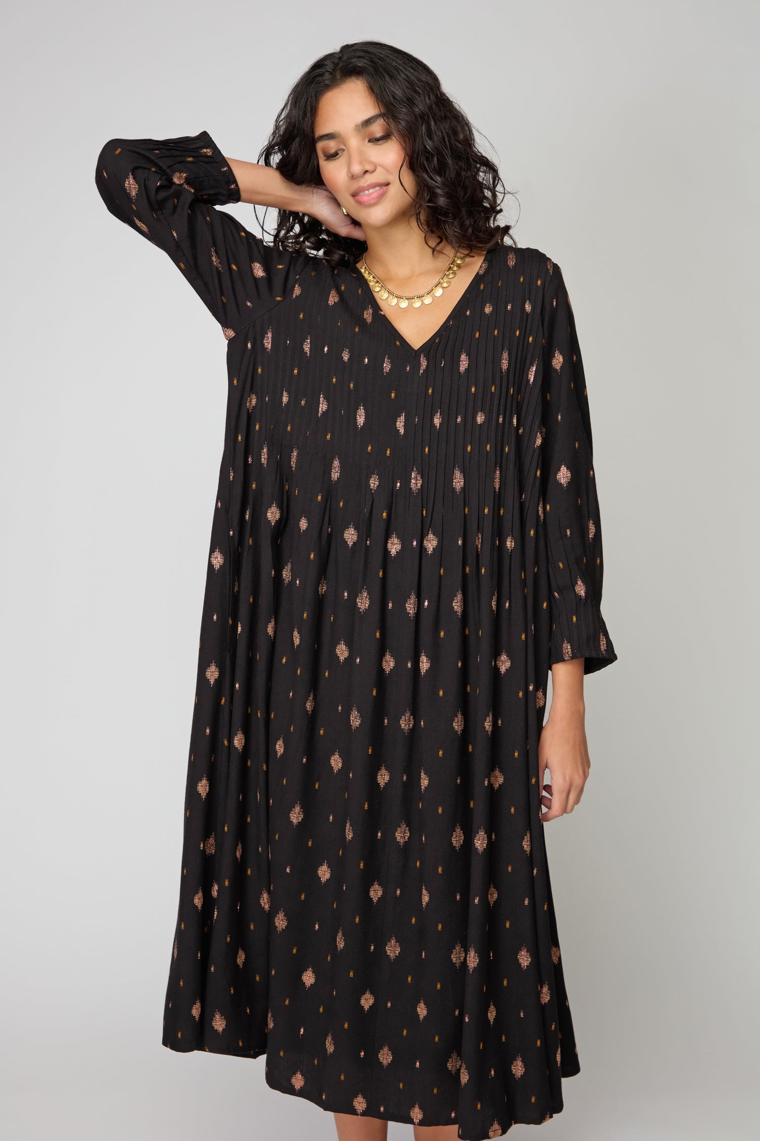 A person stands against a plain background, wearing the Diamond Lurex Pleated Dress—a loose-fitting black and gold dress with a small geometric pattern—and has one hand behind their head.
